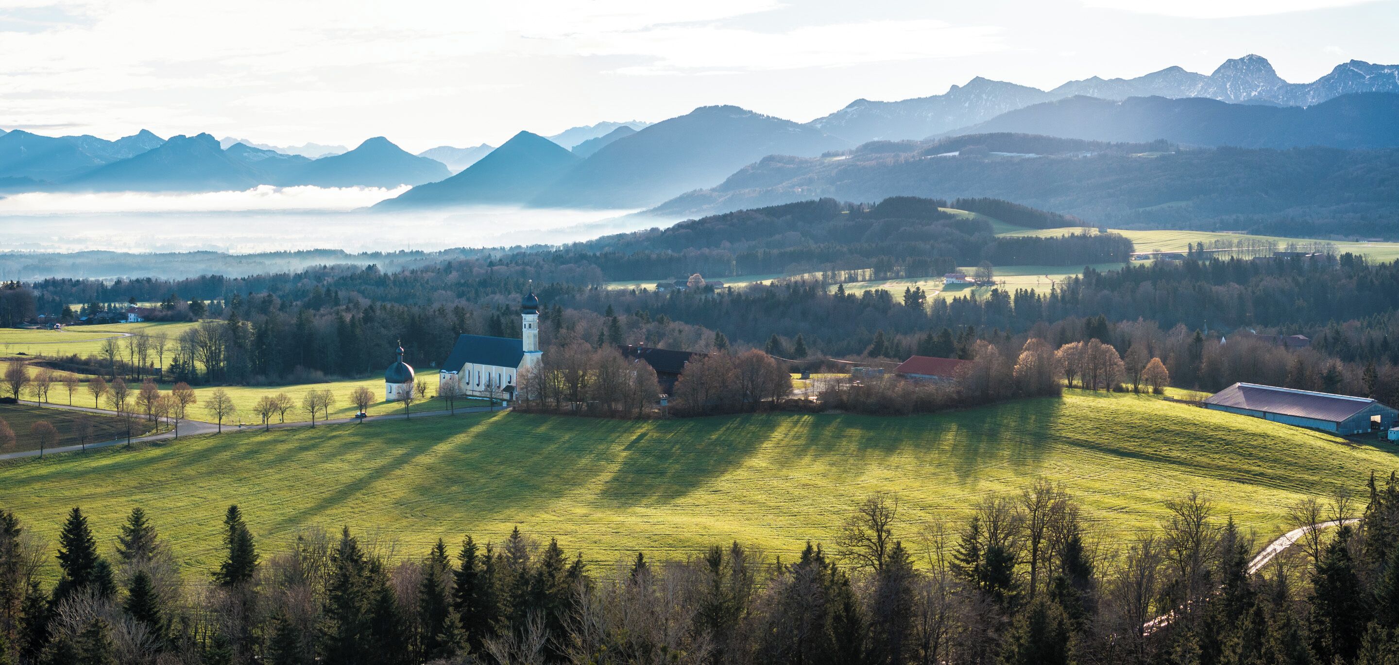 A rural landscape with a church, fields, and mountains in the background.