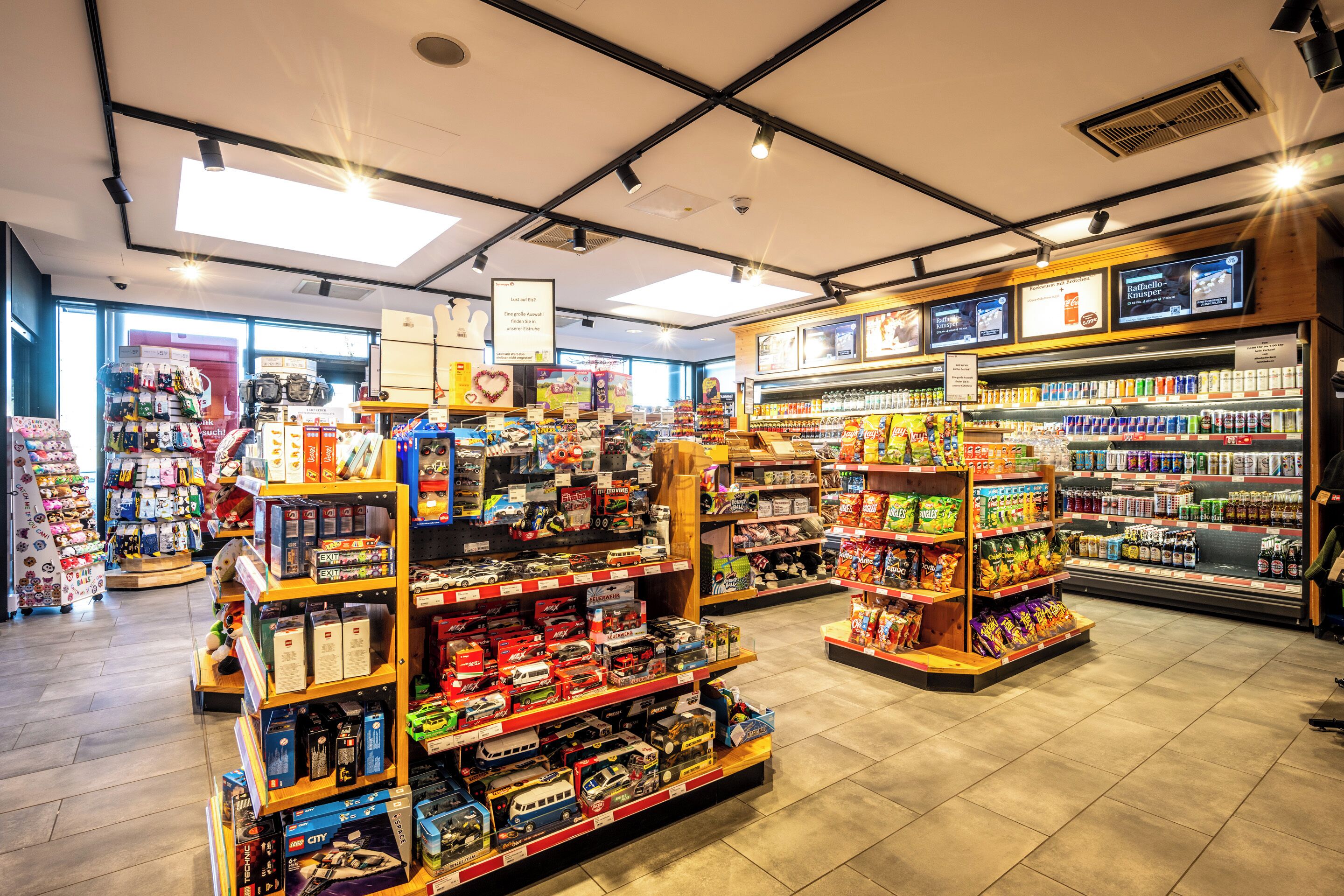 A brightly lit toy store with shelves stocked with various toys and games, a refrigerated section with snacks, and a checkout area.