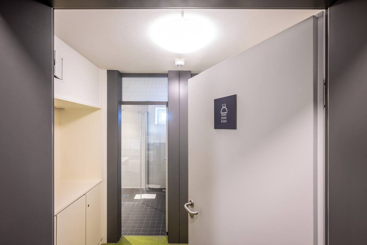 A narrow hallway with a glass door leading to a shower area, a white door with a handle, and a light fixture on the ceiling.