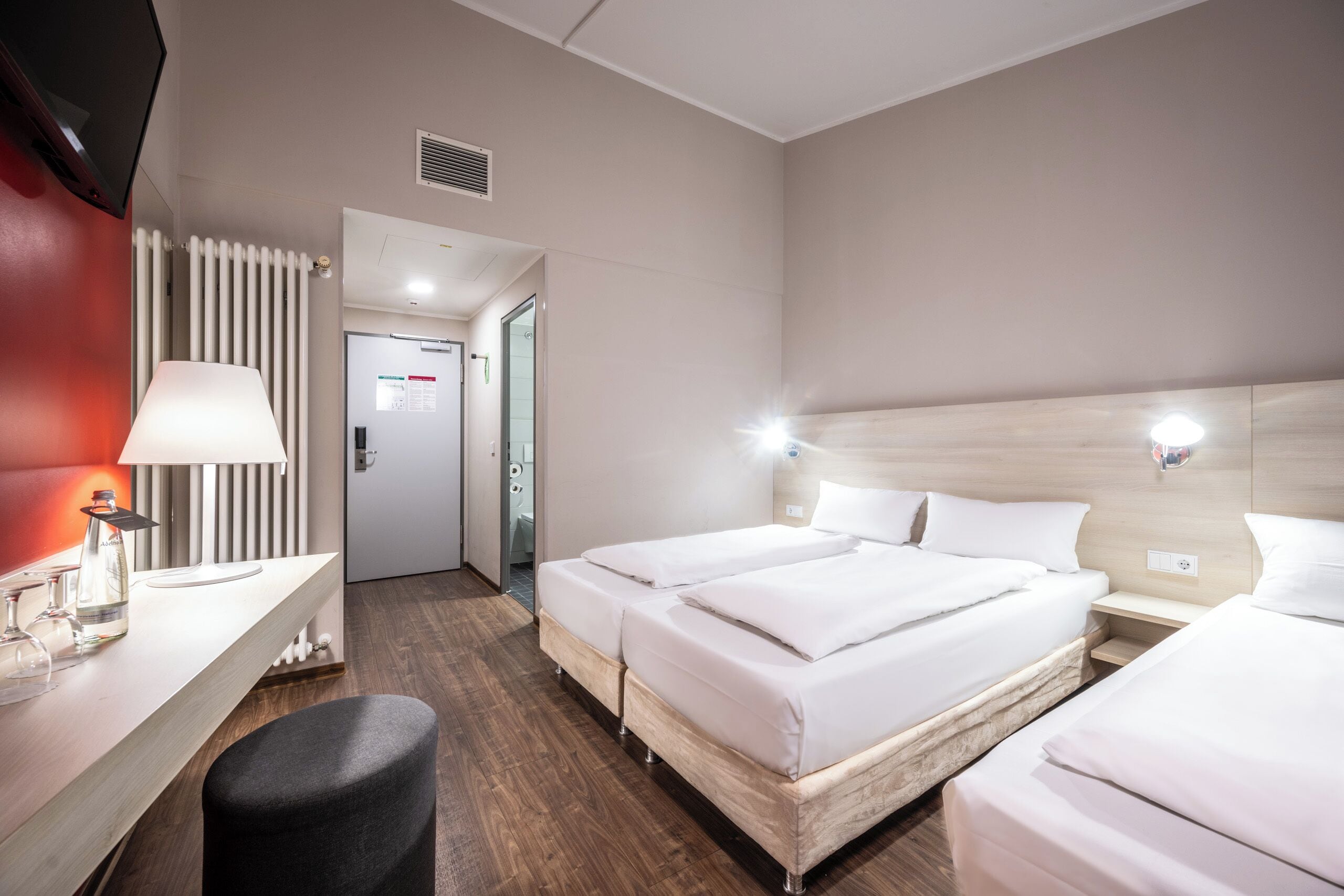 Une chambre d’hôtel avec deux lits, un bureau avec une lampe, un mur d’accent rouge et une vue sur le couloir.