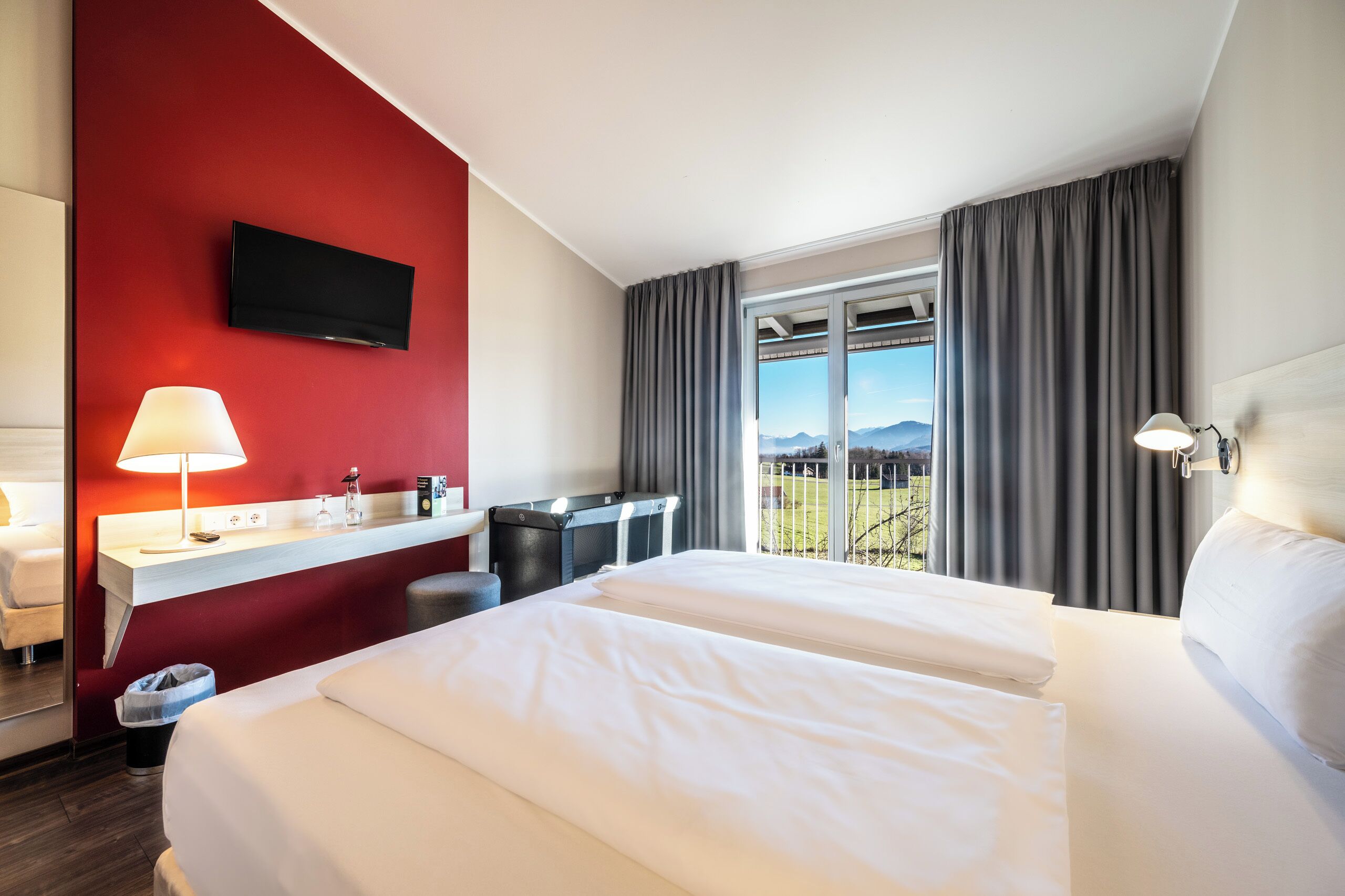 Habitación de hotel con una cama grande, un escritorio con lámpara, un televisor montado en la pared y una ventana con vistas a las montañas.