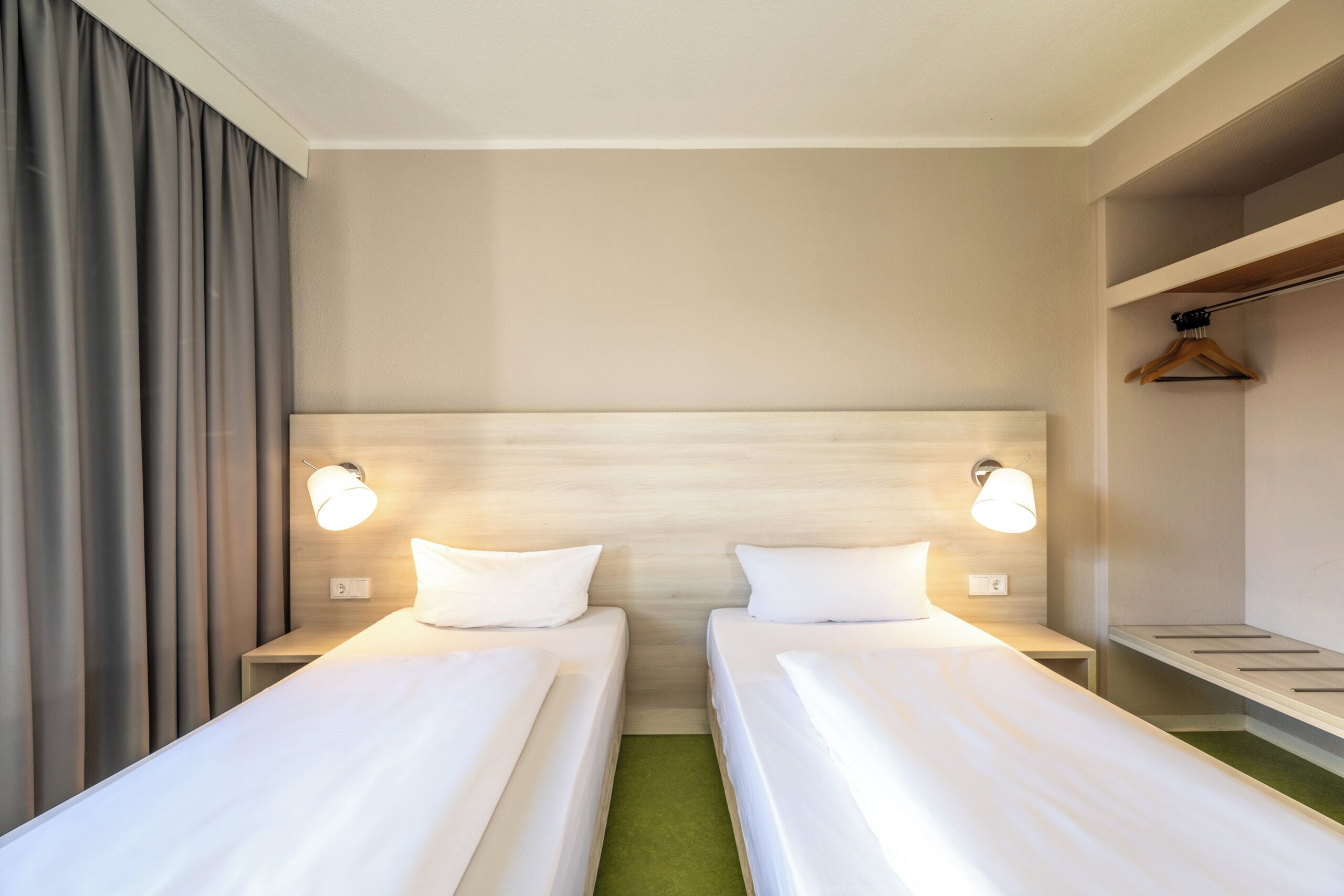 A hotel room with two single beds, a nightstand with a lamp, a wooden headboard, and a small shelf with a hanger.