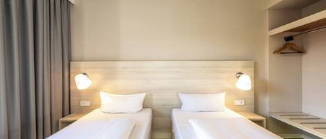 A hotel room with two single beds, a nightstand with a lamp, a wooden headboard, and a small shelf with a hanger.