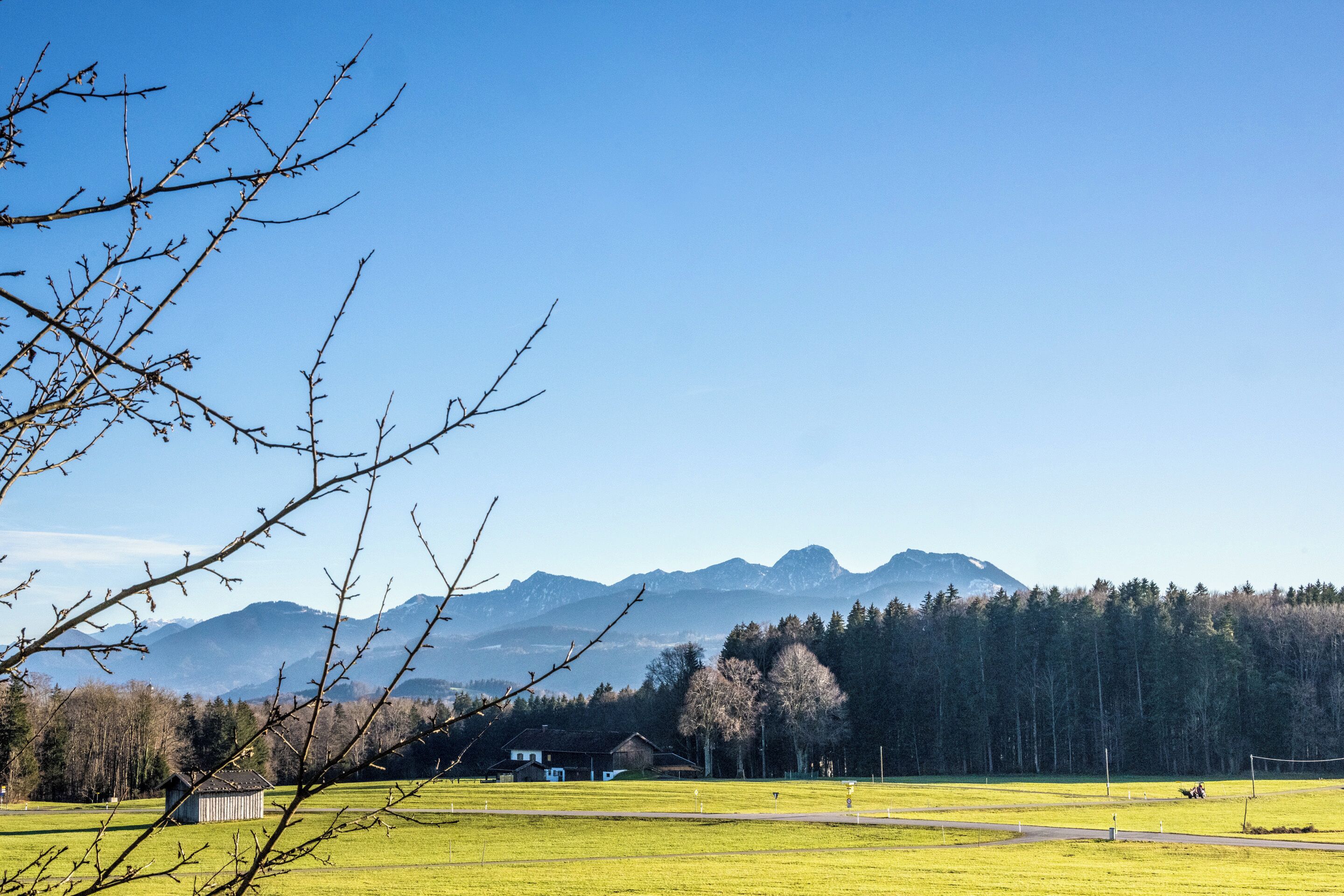 A rural landscape with a clear sky, a green field, a small building, and mountains in the background.