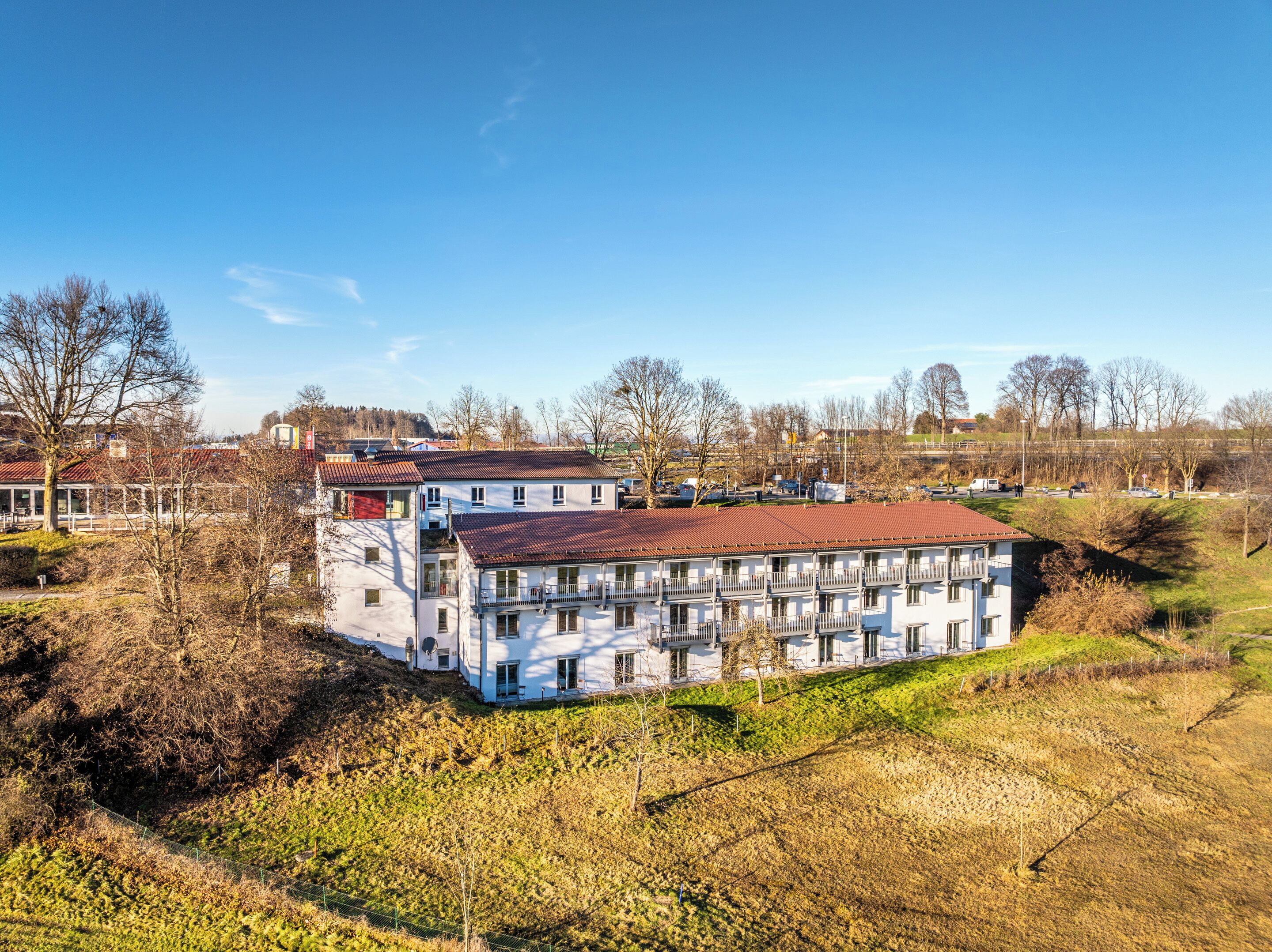 A multi-story building with balconies, surrounded by trees and a hilly landscape.