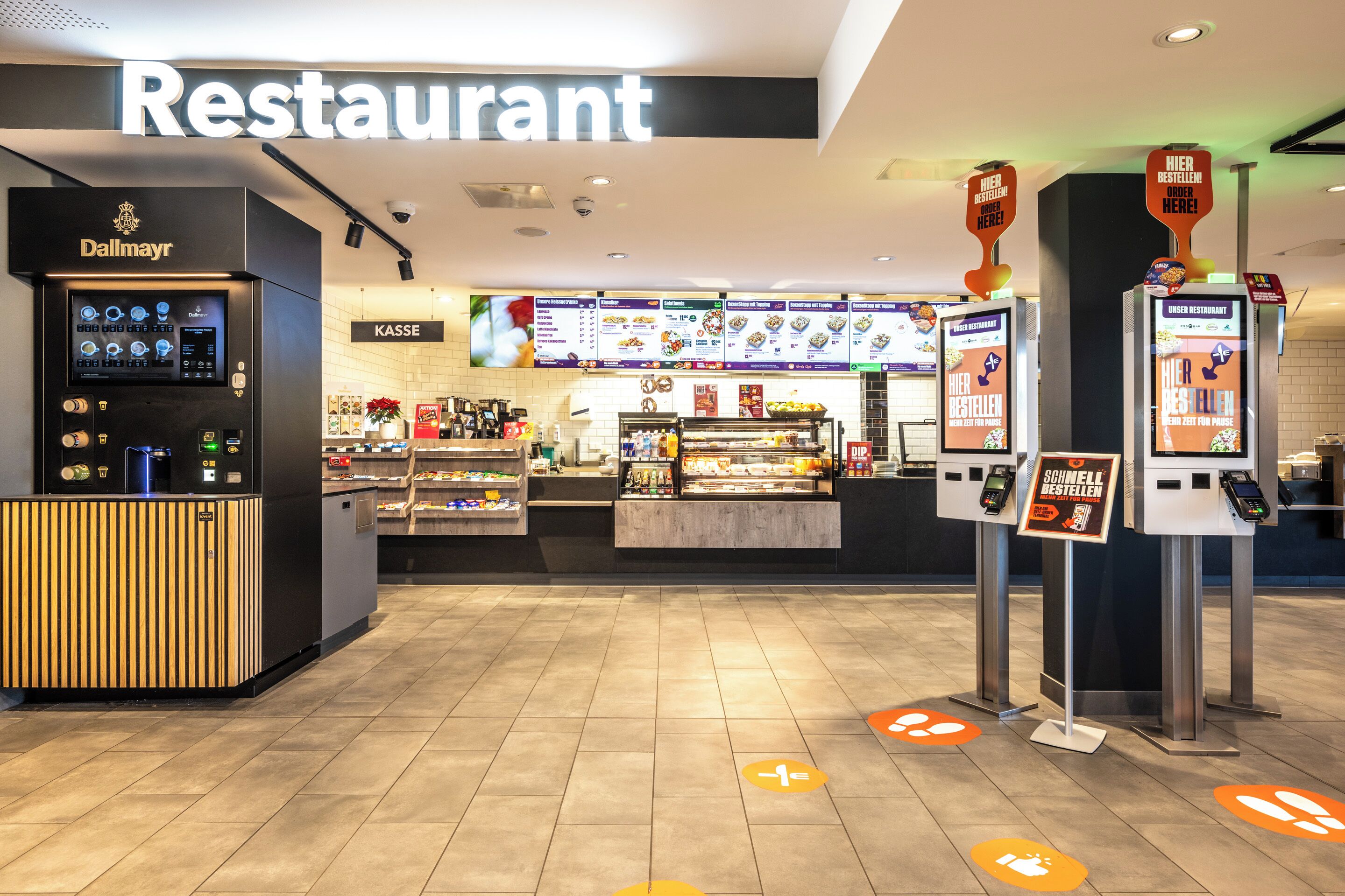 A restaurant with a self-service kiosk, a Kasse counter, and digital menu displays.