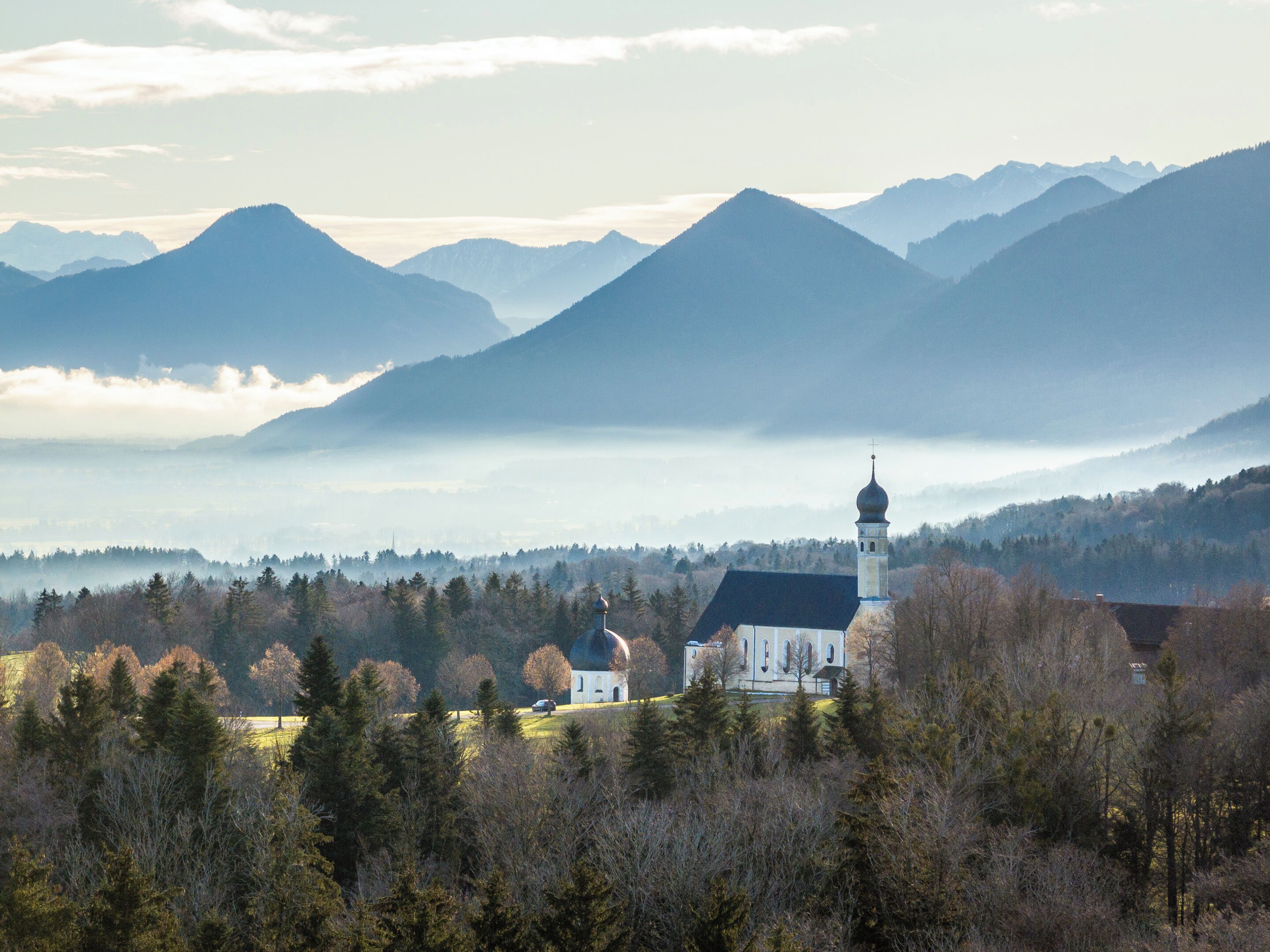A church with a bell tower surrounded by a forest and mountains in the background.
