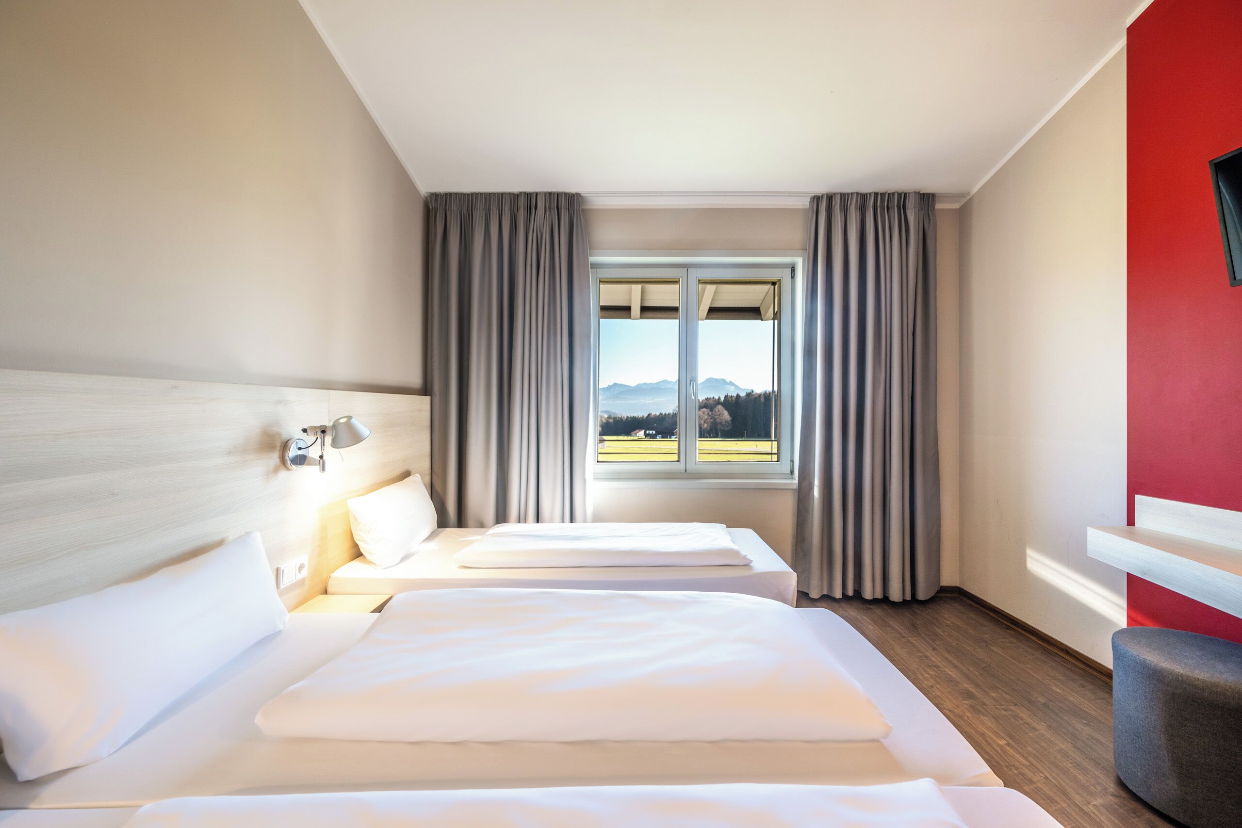 Habitación de hotel con dos camas, un ventanal con cortinas, una pared de acento rojo y un cabecero de madera.