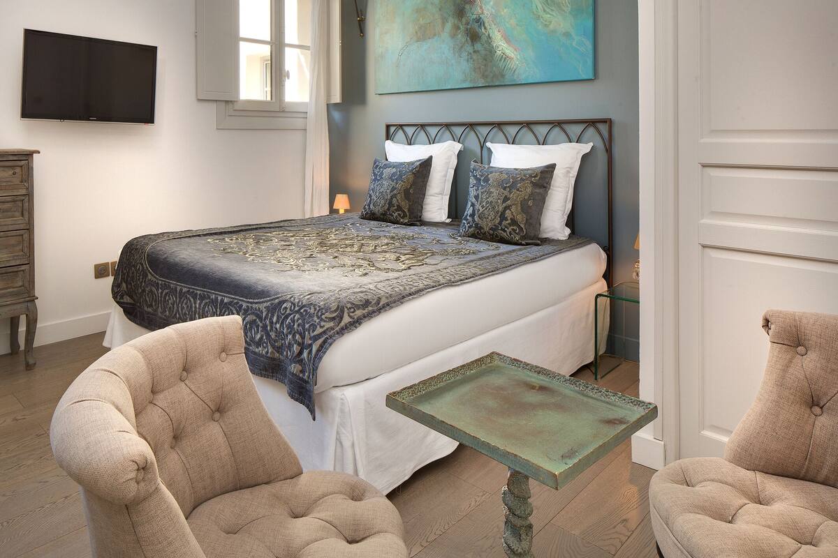 A bedroom with a bed, two armchairs, a small table, a dresser, and a painting on the wall.