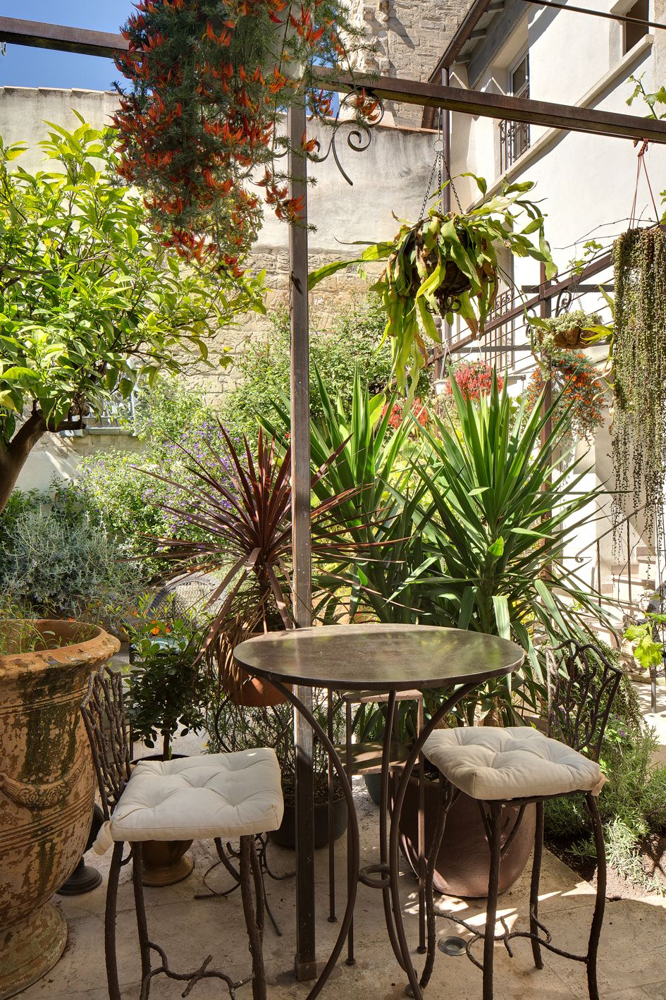 Une terrasse avec une table ronde et deux chaises, entourée de plantes en pot et d’une plante suspendue.