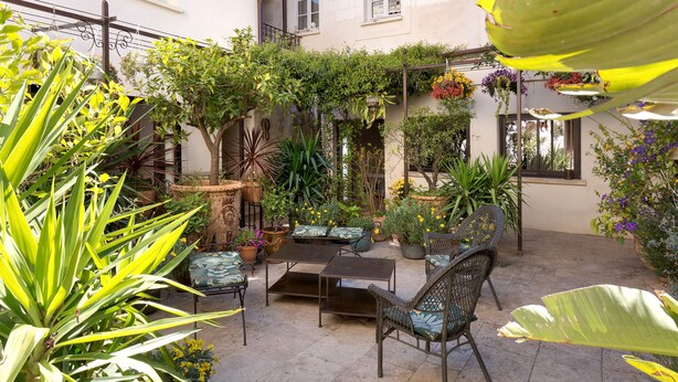 Une cour avec une table et des chaises, entourée de plantes en pot et de suspensions fleuries.