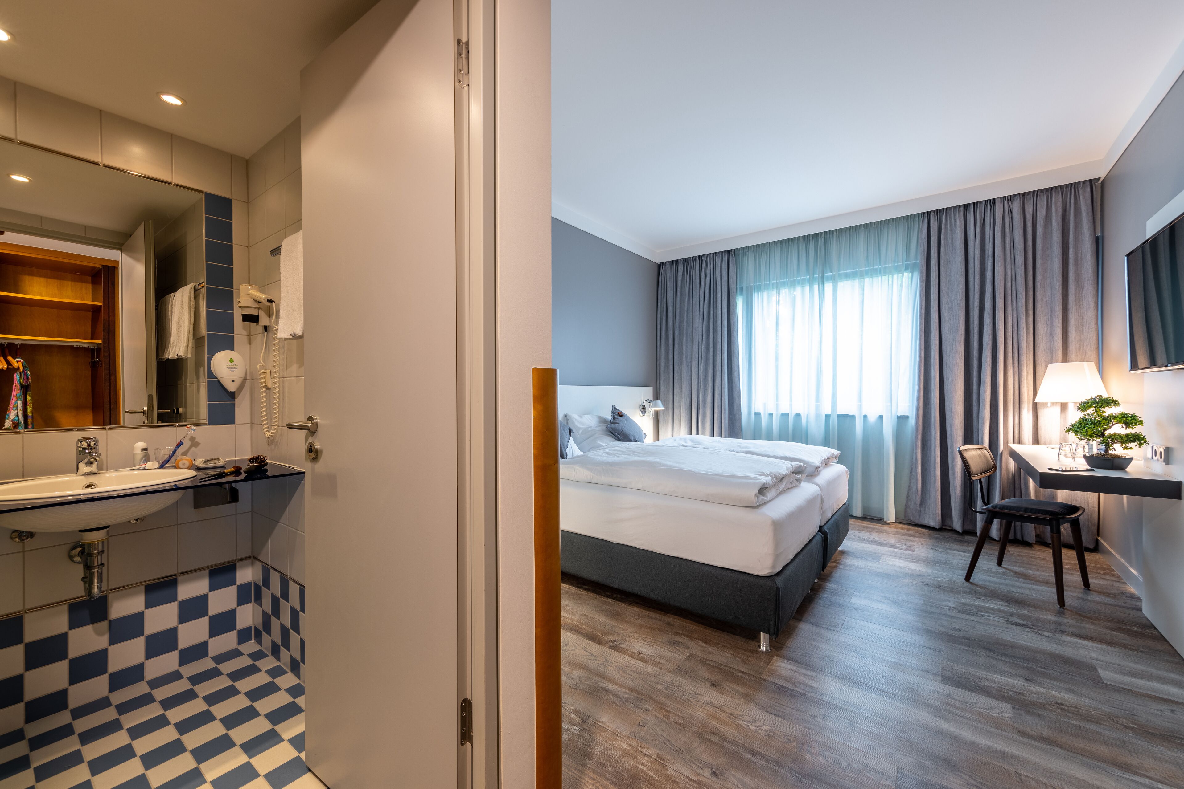 A hotel room with a checkered floor, a bed, a desk with a lamp, and a large window with curtains.