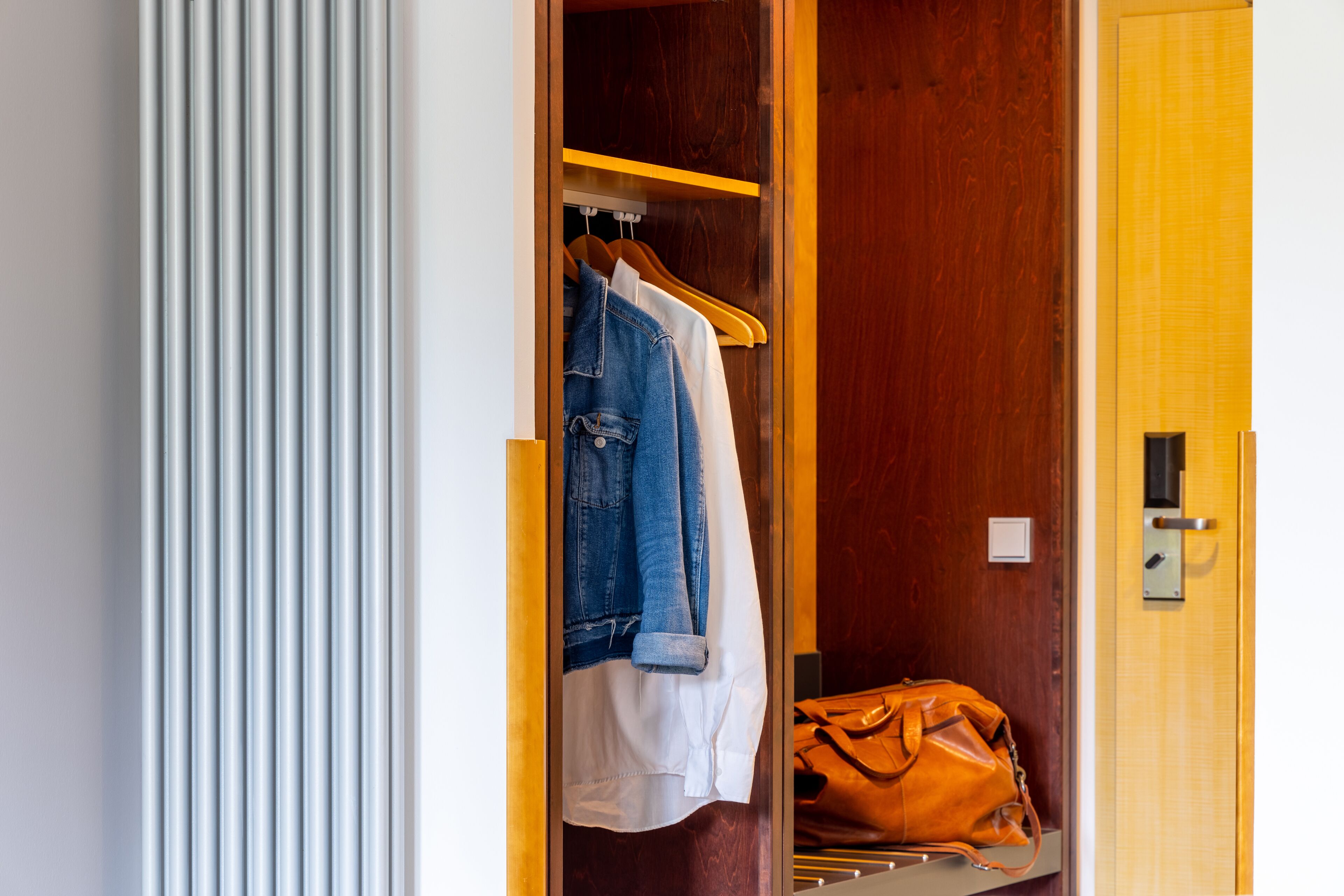 A hotel room with a wooden wardrobe, a hanging garment, and a handbag on the floor.