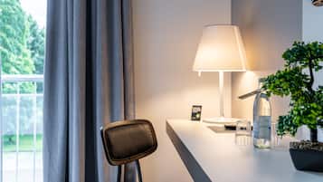 A modern hotel room with a desk, a lamp, a potted plant, and a view of greenery outside.