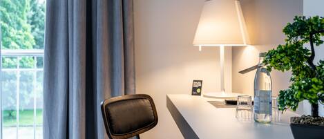 Une chambre d’hôtel moderne, équipée d’un bureau, d’une lampe, d’une plante en pot et offrant une vue sur la verdure environnante.