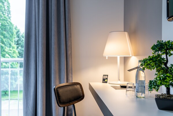 Une chambre d’hôtel moderne, équipée d’un bureau, d’une lampe, d’une plante en pot et offrant une vue sur la verdure environnante.