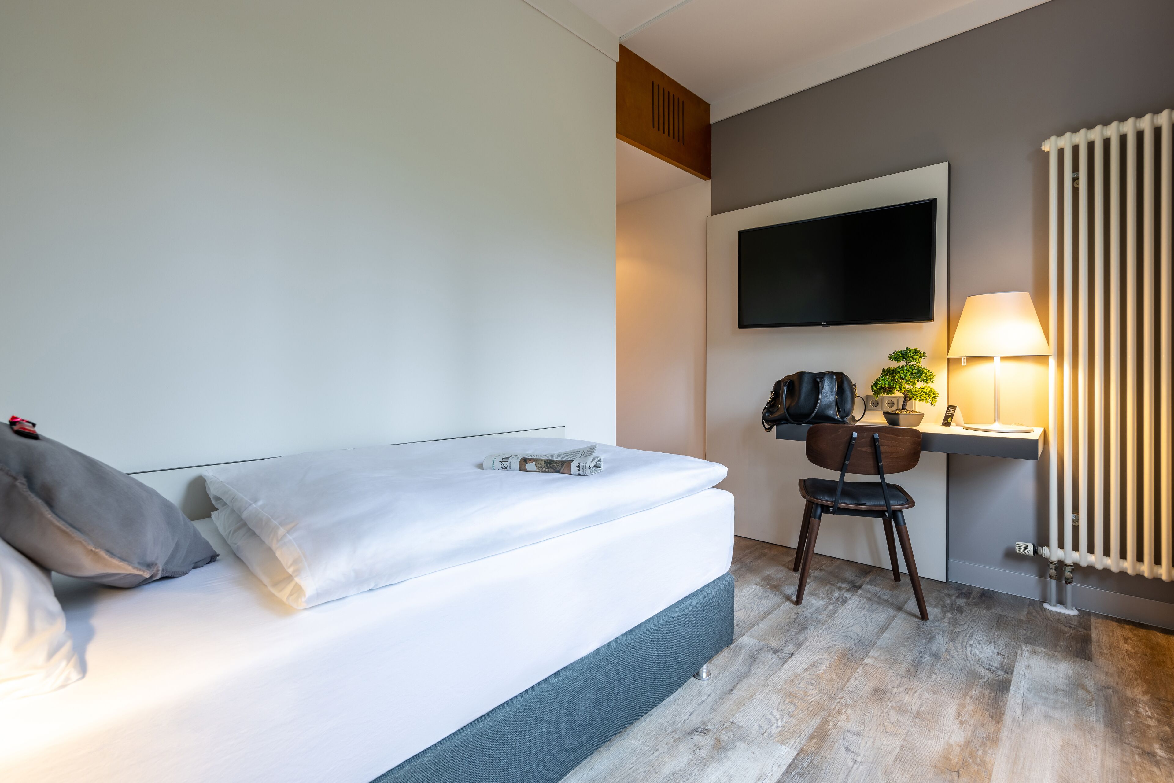 A modern hotel room with a bed, a desk with a chair, a TV mounted on the wall, and a radiator.
