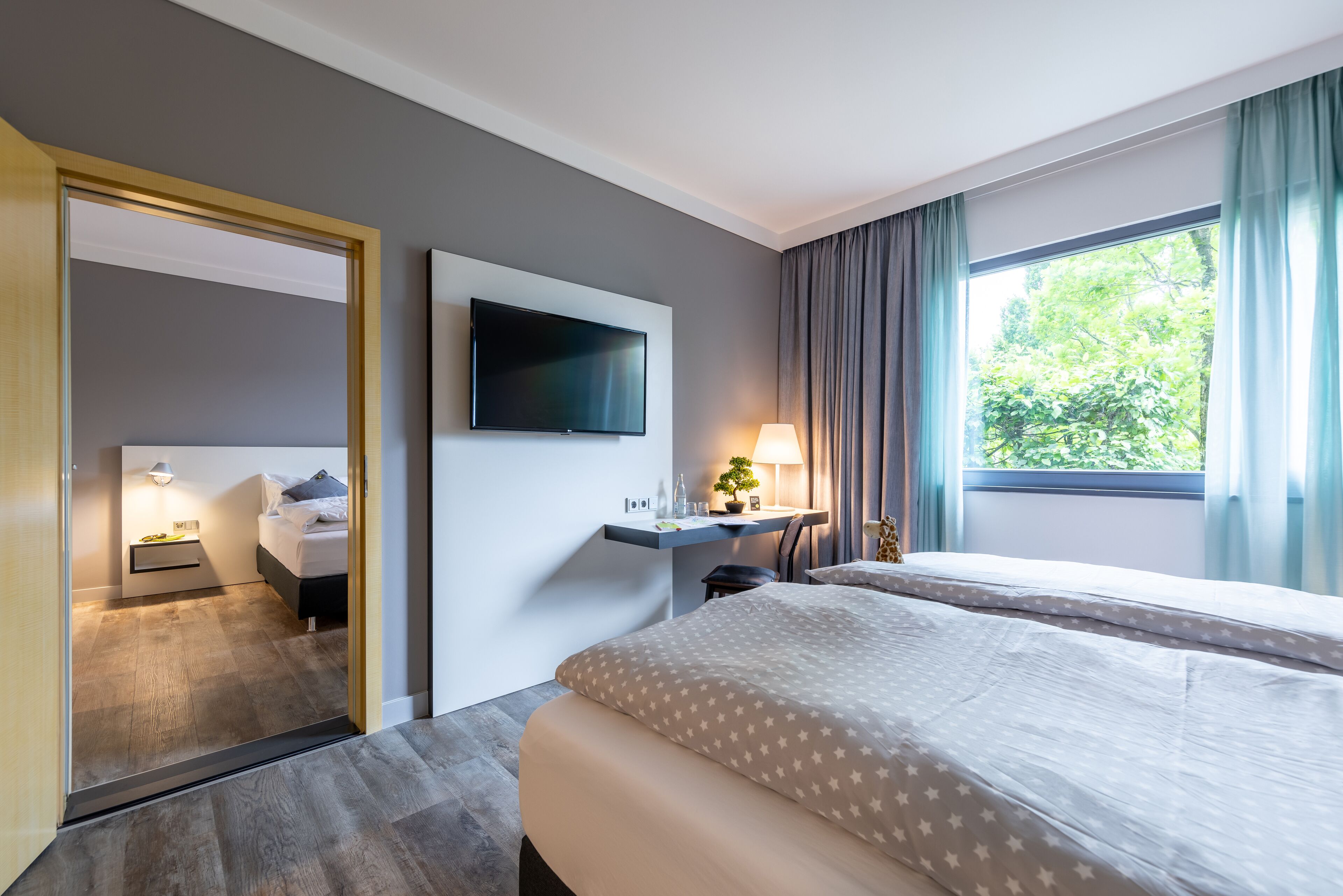 A hotel room with a bed, a desk with a lamp, a TV mounted on the wall, and a window with curtains.