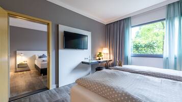 A hotel room with a bed, a desk with a lamp, a TV mounted on the wall, and a window with curtains.