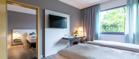 A hotel room with a bed, a desk with a lamp, a TV mounted on the wall, and a window with curtains.