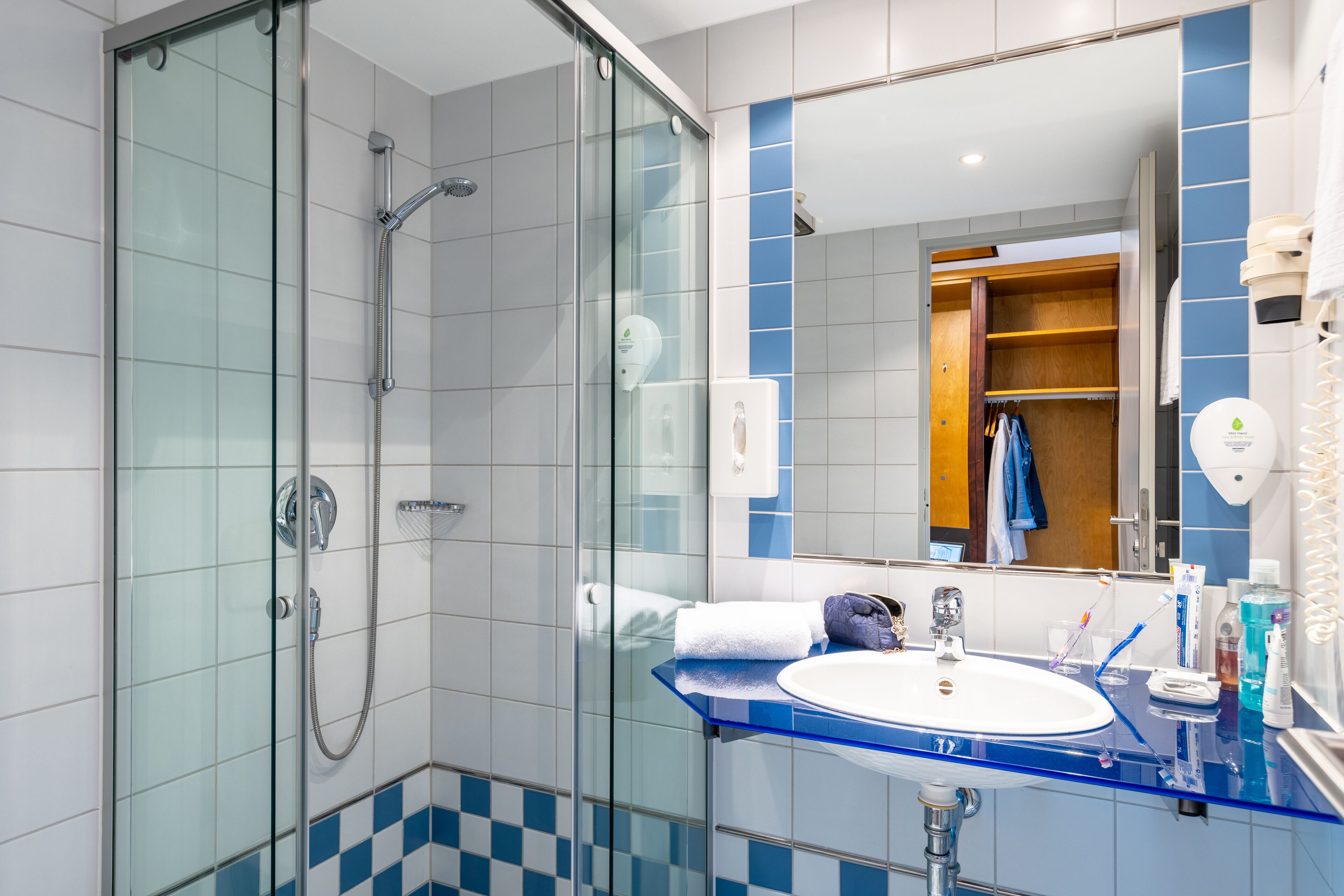 Une salle de bain moderne avec une douche à l’italienne en verre, un meuble-lavabo aux carreaux bleus et un grand miroir qui reflète un placard en bois.
