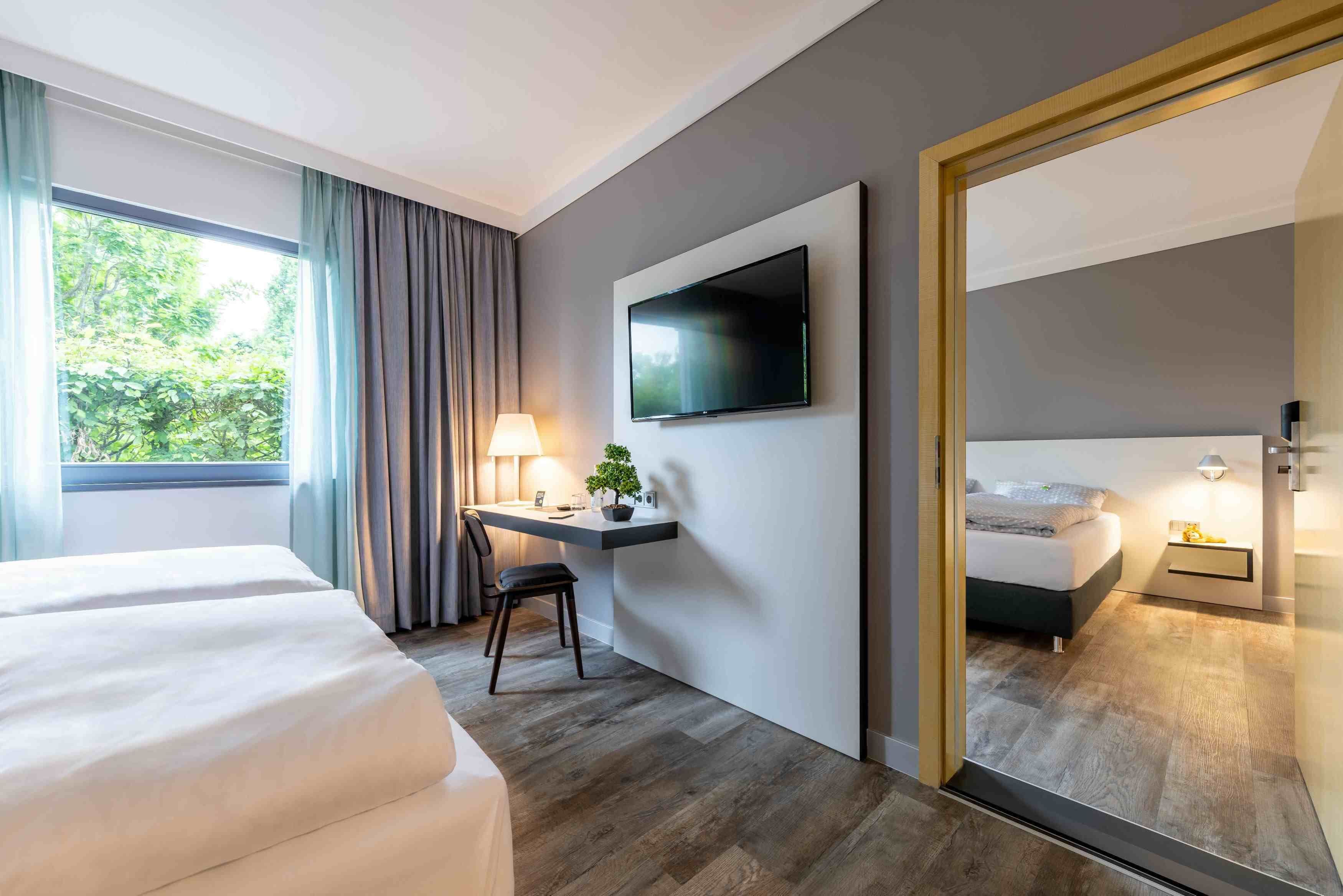 A hotel room with a bed, a desk with a chair, a TV mounted on the wall, and a large window with curtains.