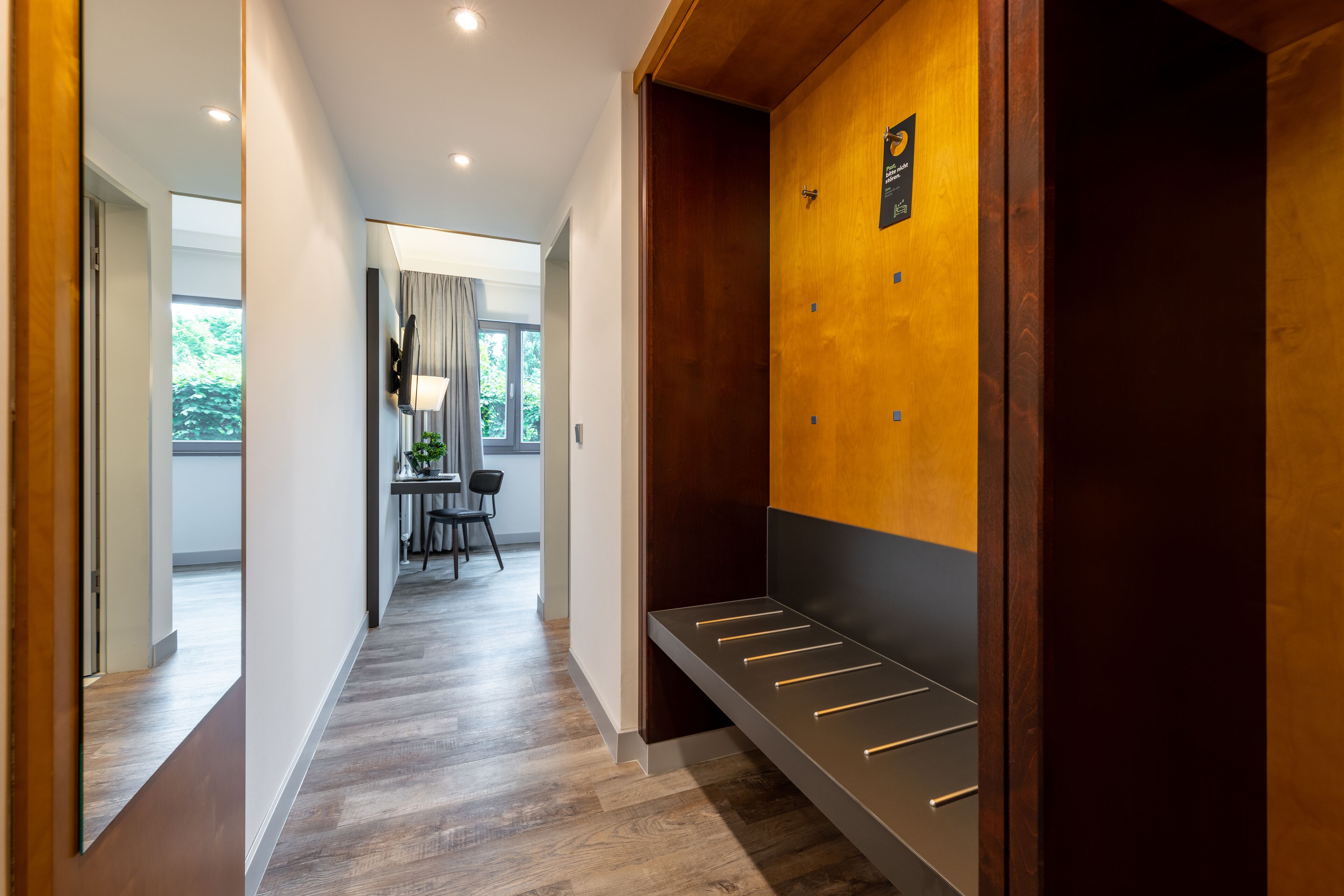 A modern hotel hallway with a wooden bench, a coat rack, and a television mounted on the wall.