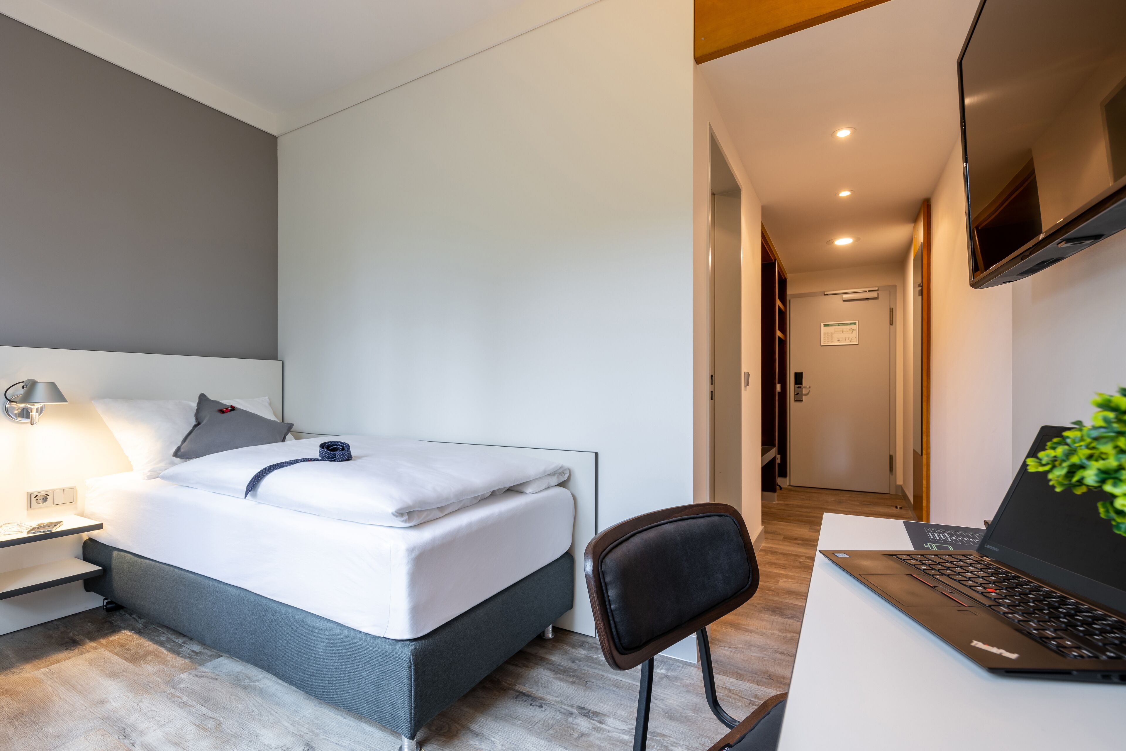 A modern hotel room with a bed, bedside table, lamp, laptop, and a TV mounted on the wall.