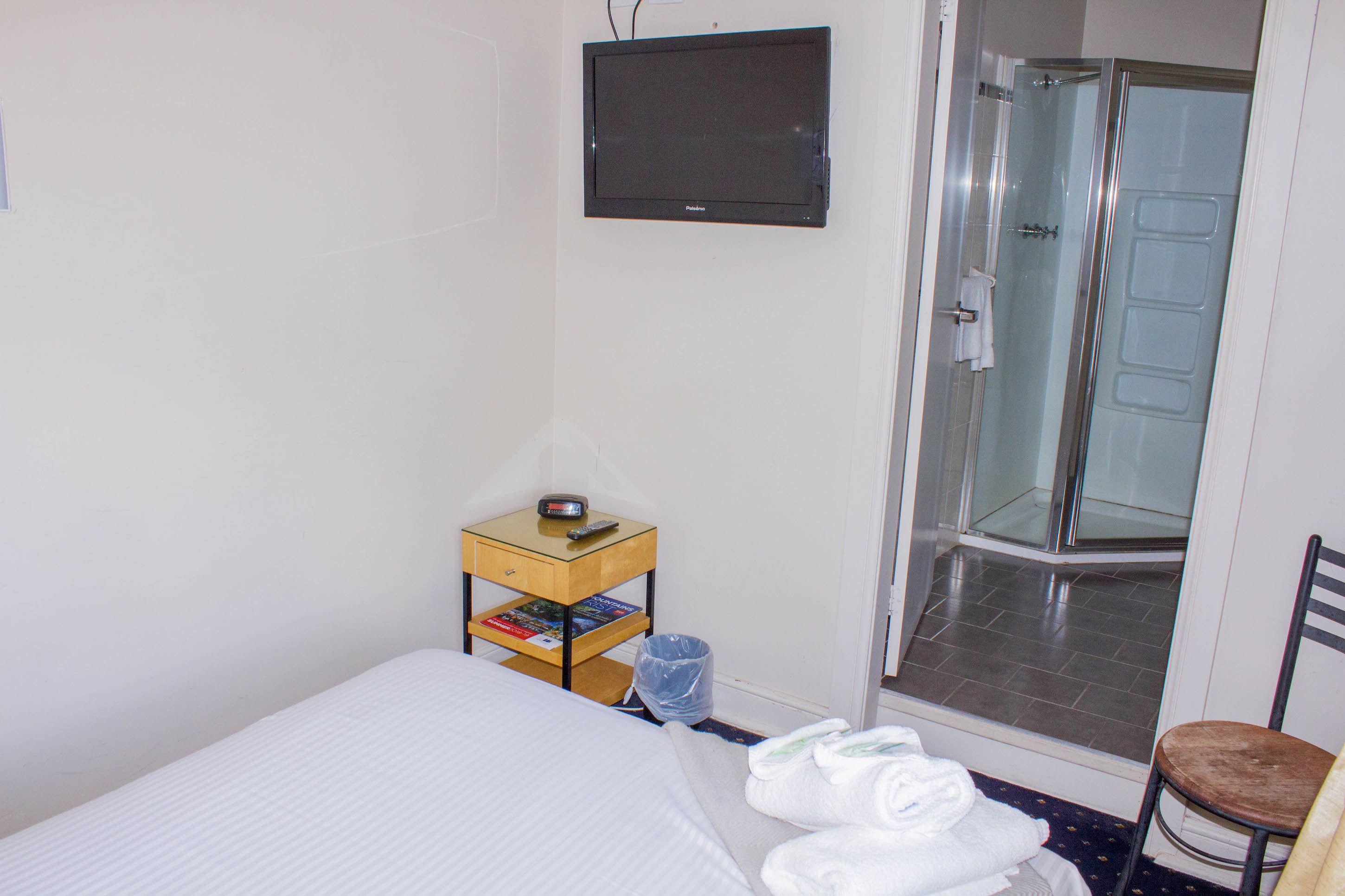 Hotelkamer met een bed, een klein houten nachtkastje met een afstandsbediening, een douchecabine van glas en een stoel.
