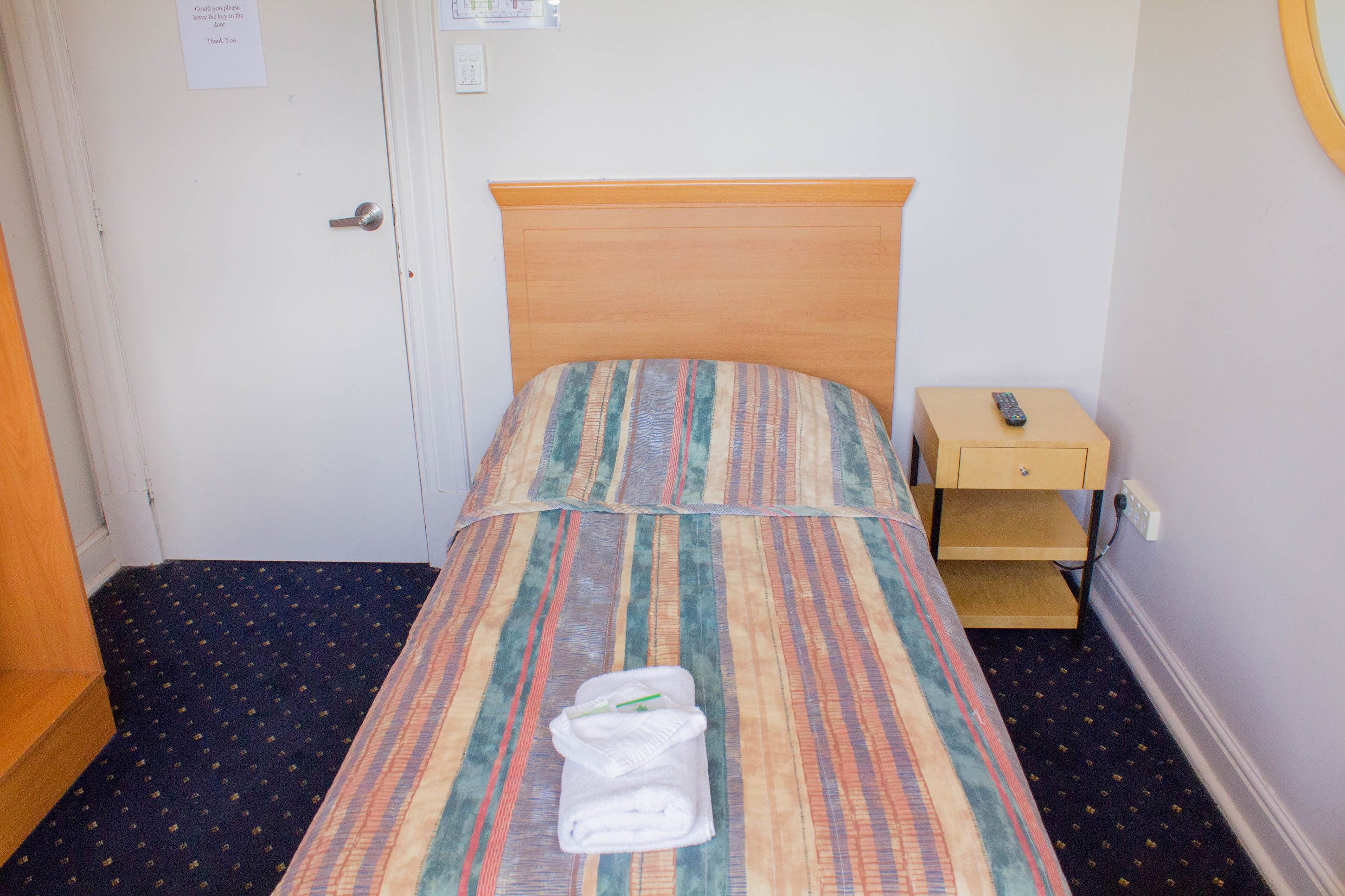 A small, single-bed room with a striped bedspread, a bedside table with a remote control, and a folded towel on the bed.