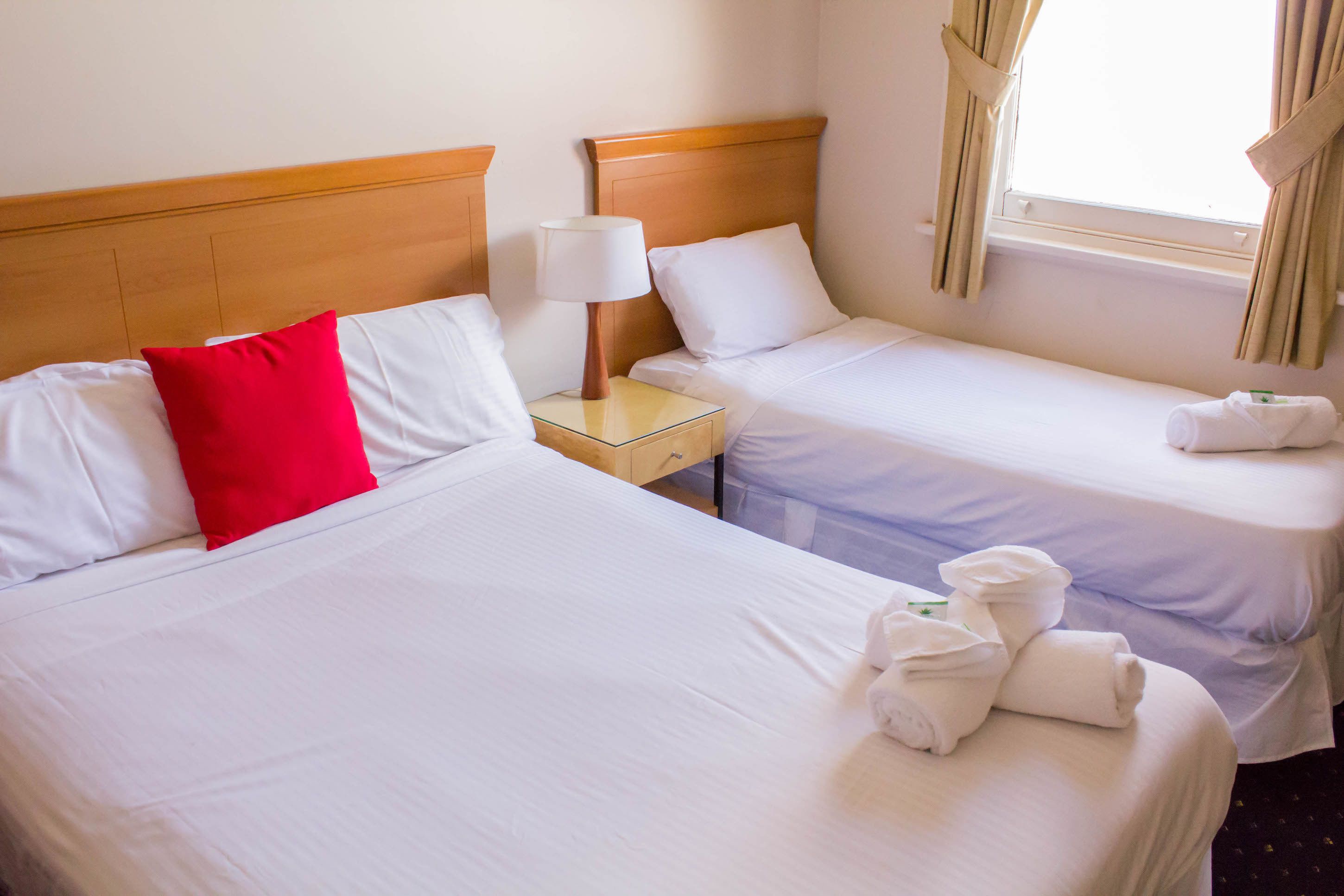Two single beds with white linens, a red pillow, a nightstand with a lamp, and a window with curtains.