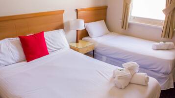 Two single beds with white linens, a red pillow, a nightstand with a lamp, and a window with curtains.