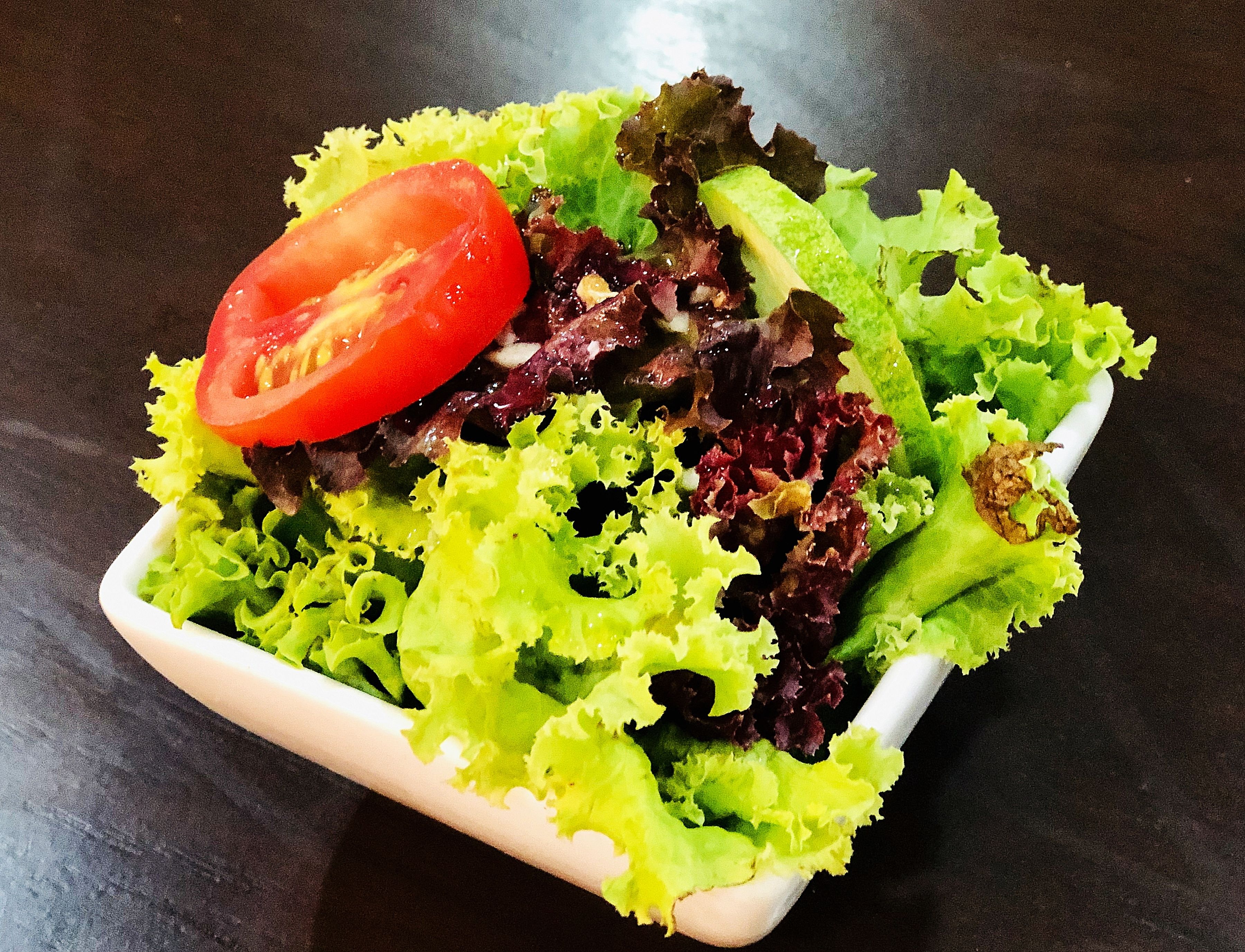 A square white bowl containing a salad with mixed green and red lettuce, and a slice of tomato on top.