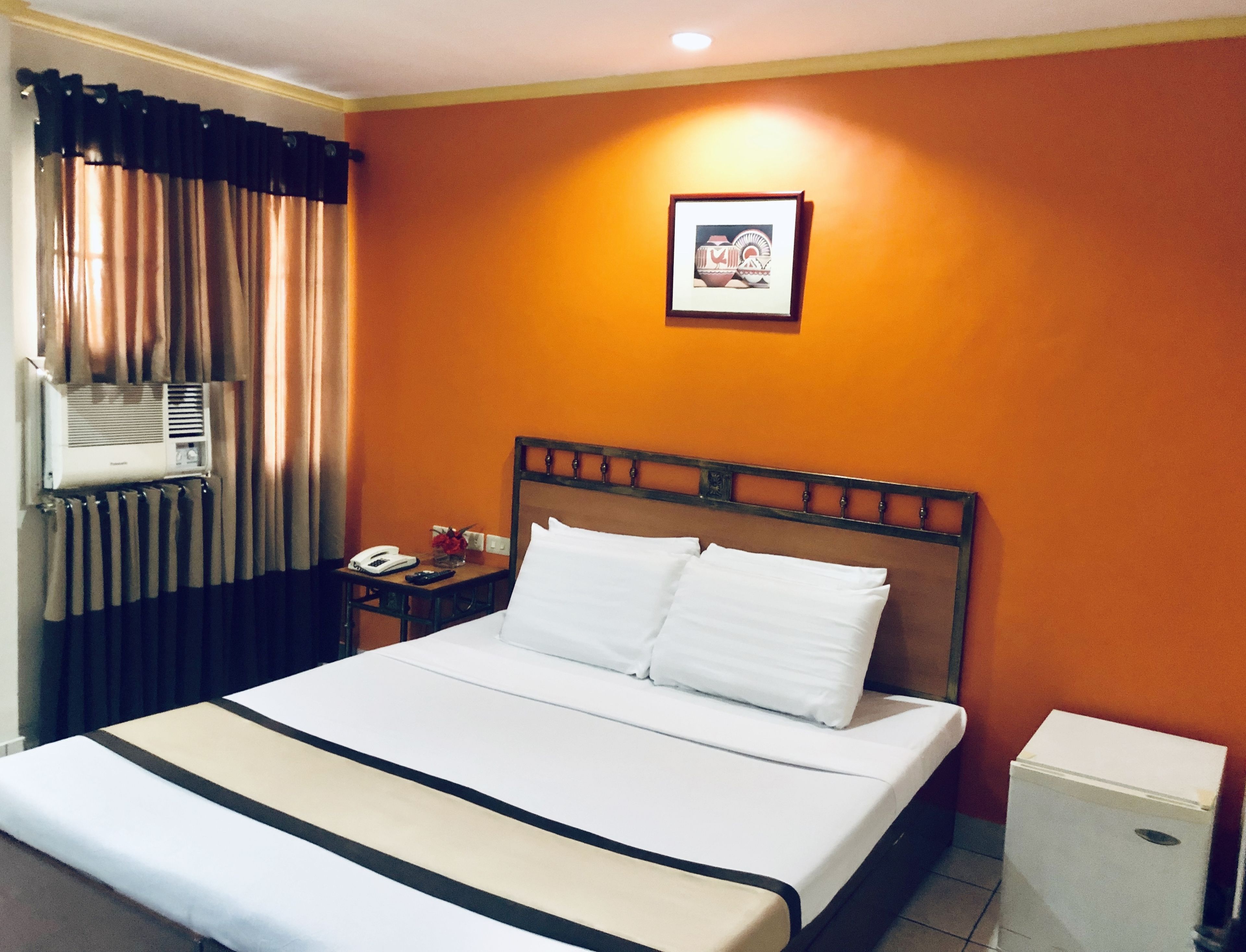 Habitación de hotel con cama, mesita de noche, teléfono, un cuadro en la pared y un equipo de aire acondicionado.