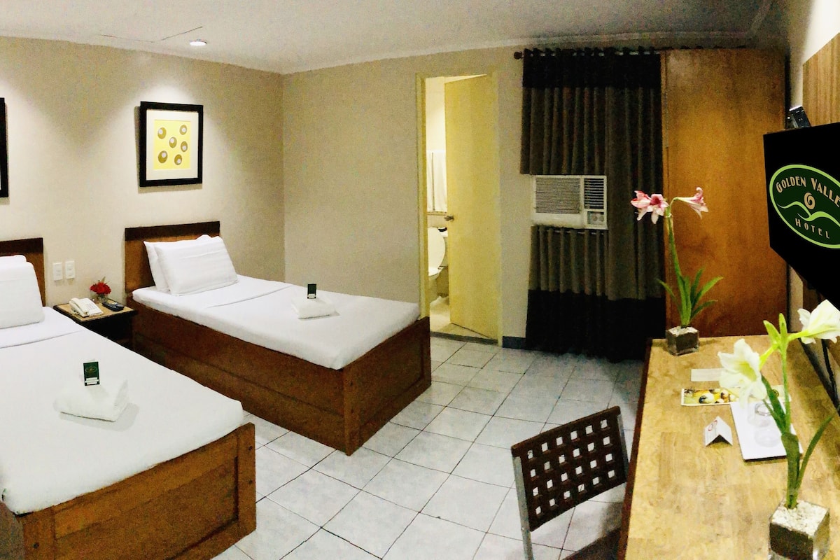 A hotel room with two single beds, a wooden headboard, a bedside table with a lamp, and a large mirror reflecting the room.