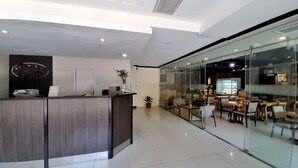 Um balcão de recepção moderno com acabamento em madeira, um vaso de planta e uma divisória de vidro separando a área da recepção do espaço de refeições.