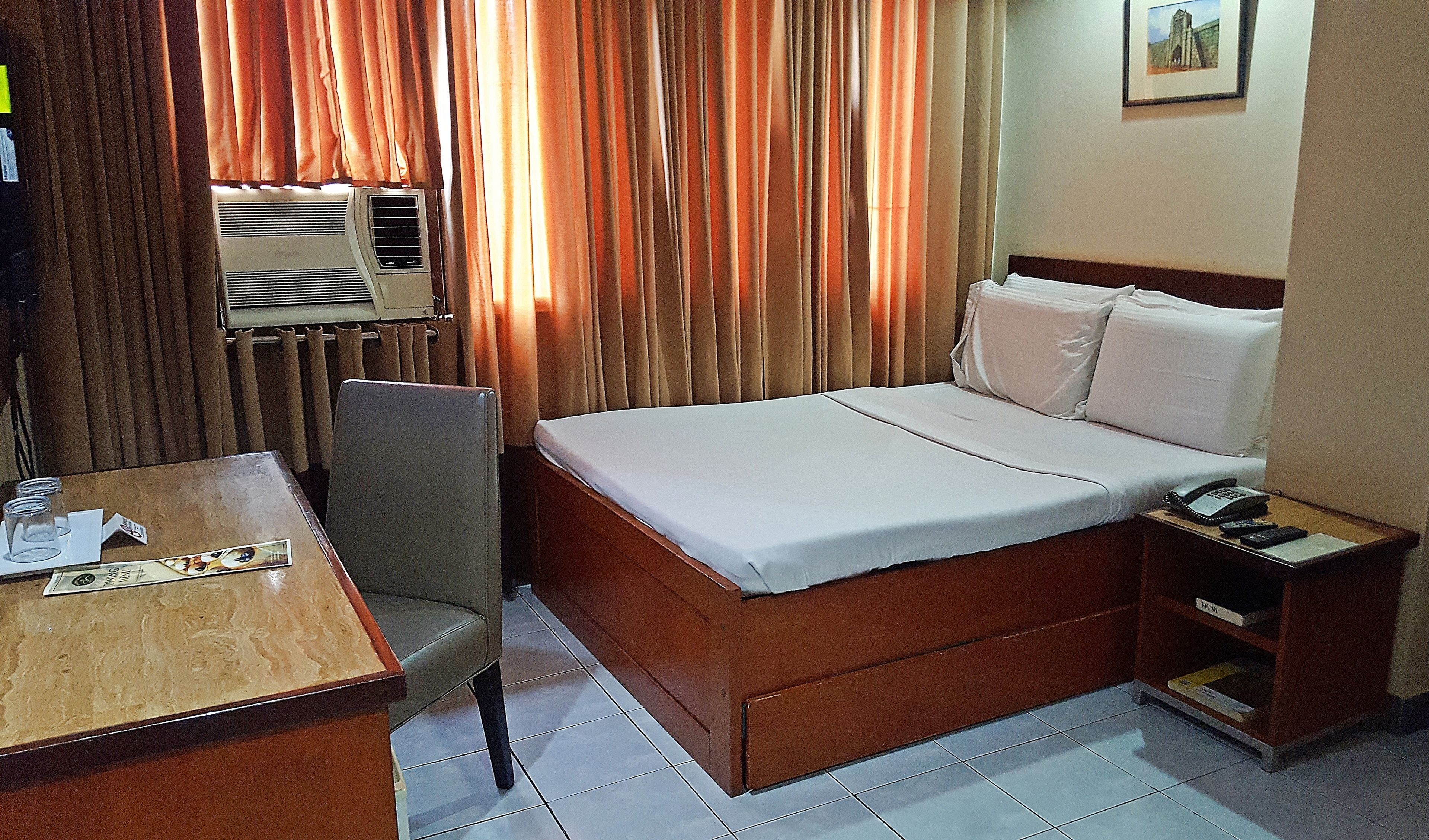A hotel room with a bed, a chair, a table with a phone and remote controls, and a window with curtains.