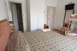A bedroom with a patterned bedspread, a wooden headboard, a small bedside table, a chair, a wardrobe, and a mirror on the wall.