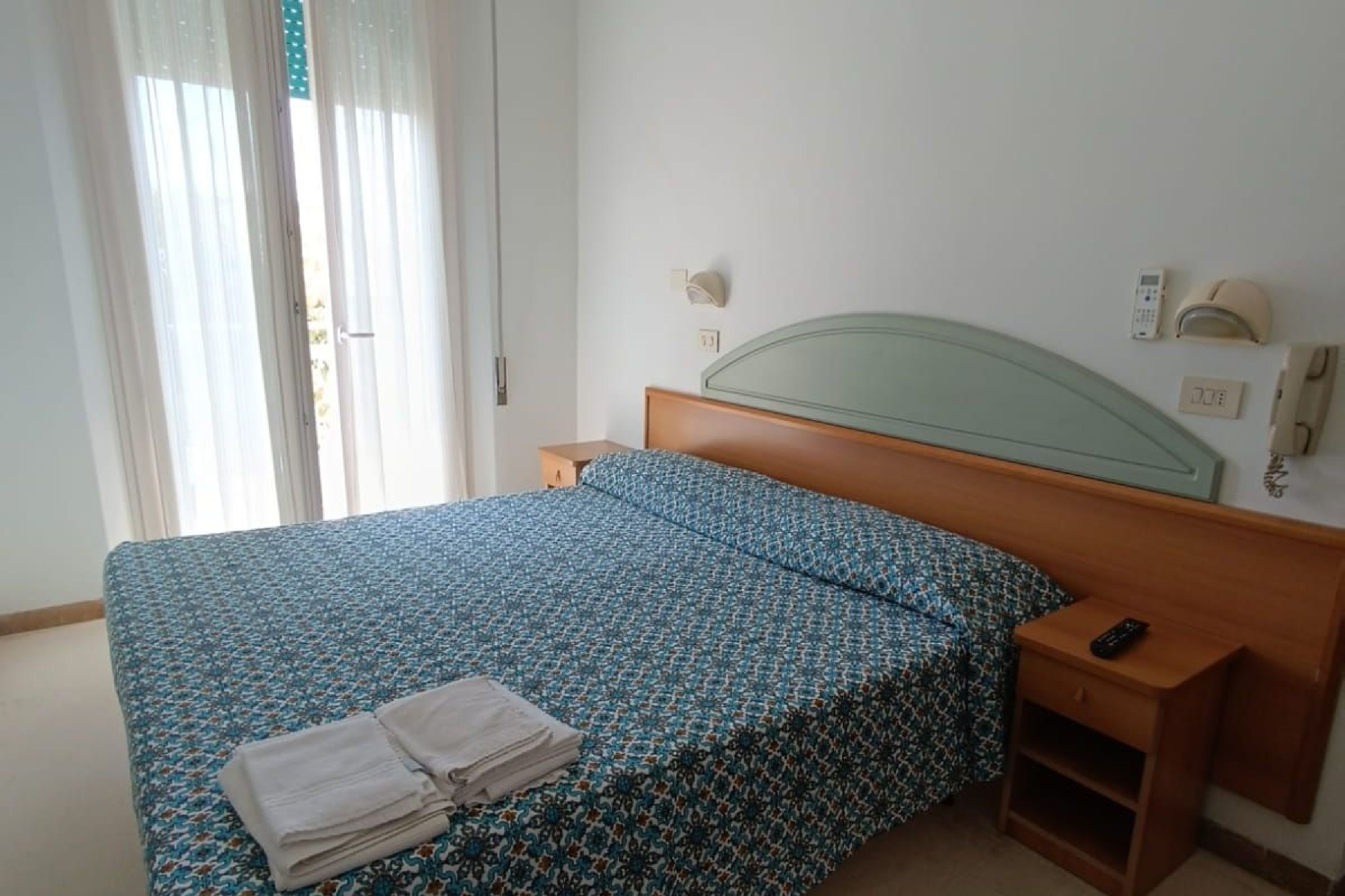 A hotel room with a bed, a nightstand, a remote control, and a window with curtains.