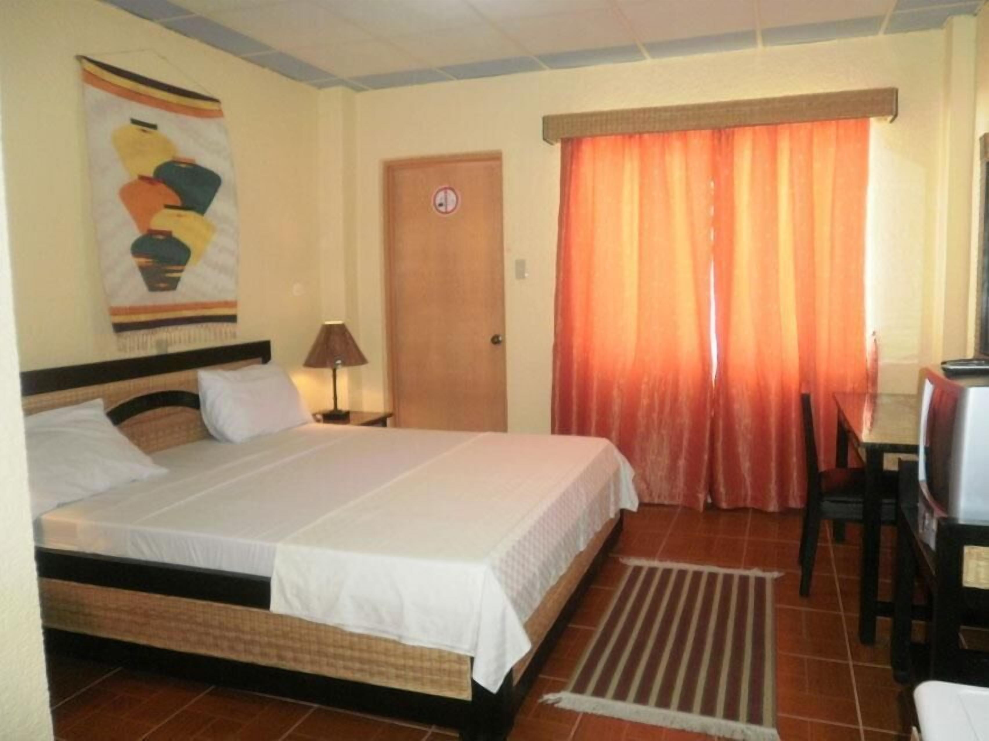 A hotel room with a bed, a desk, a chair, a lamp, and a door.