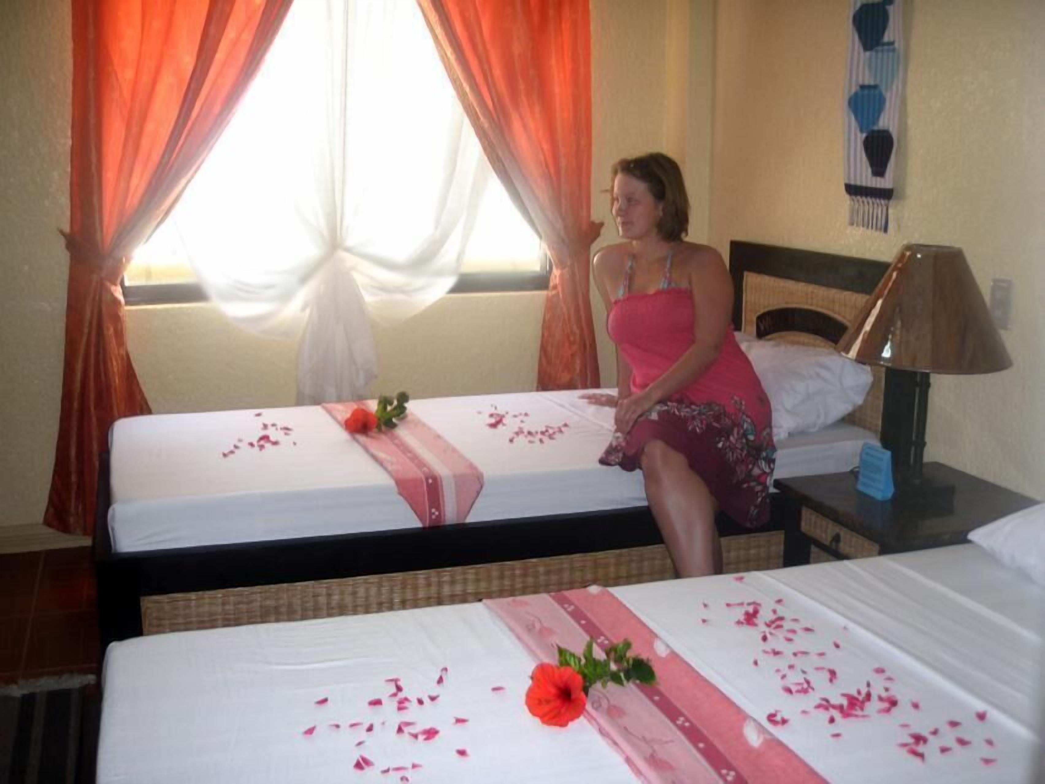 A hotel room with two beds, a nightstand with a lamp, and a window with orange curtains.