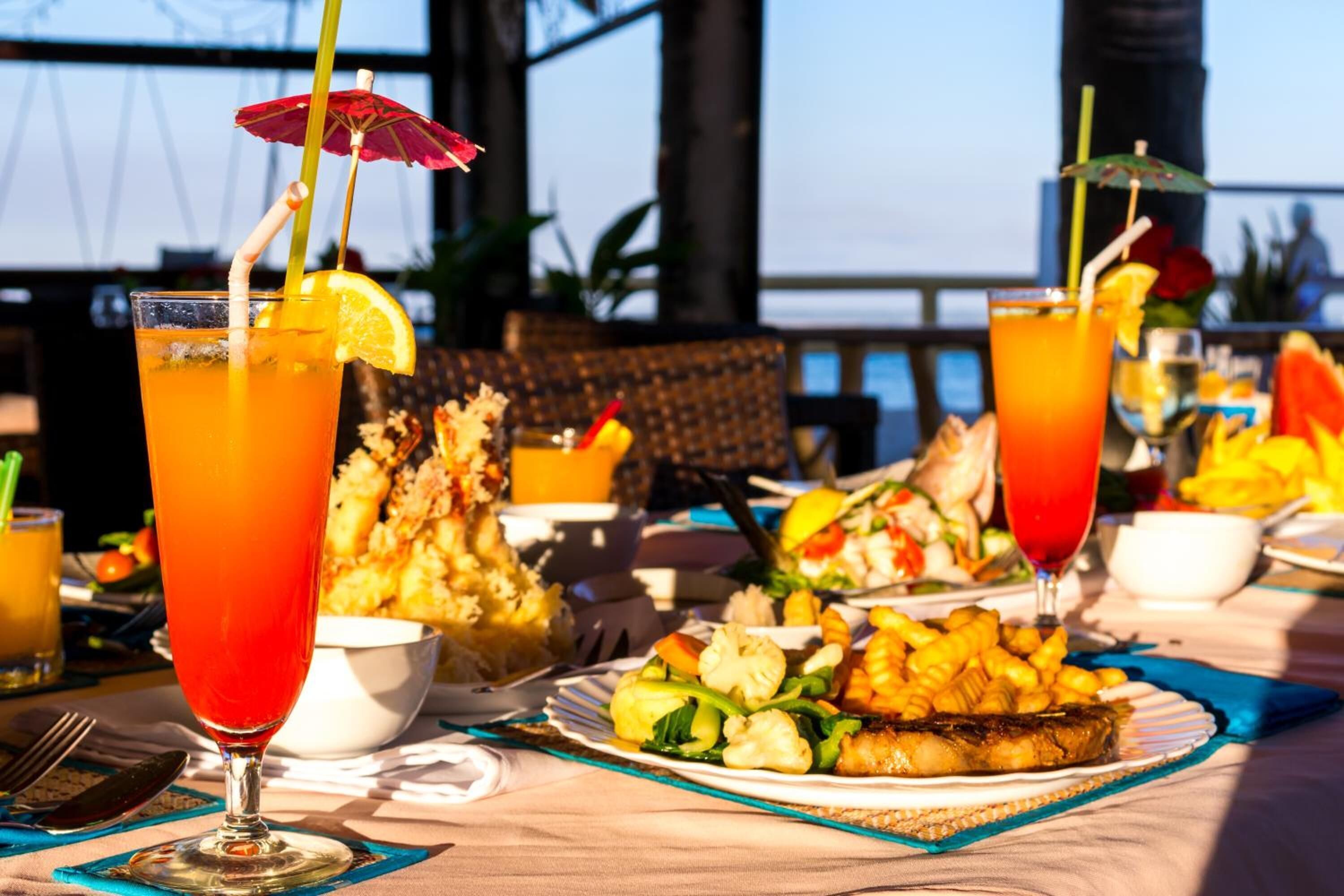 Una mesa dispuesta con diversos platos, incluyendo un filete de pescado y una ensalada, junto con dos vasos altos de una bebida roja con pajitas y una rodaja de naranja.