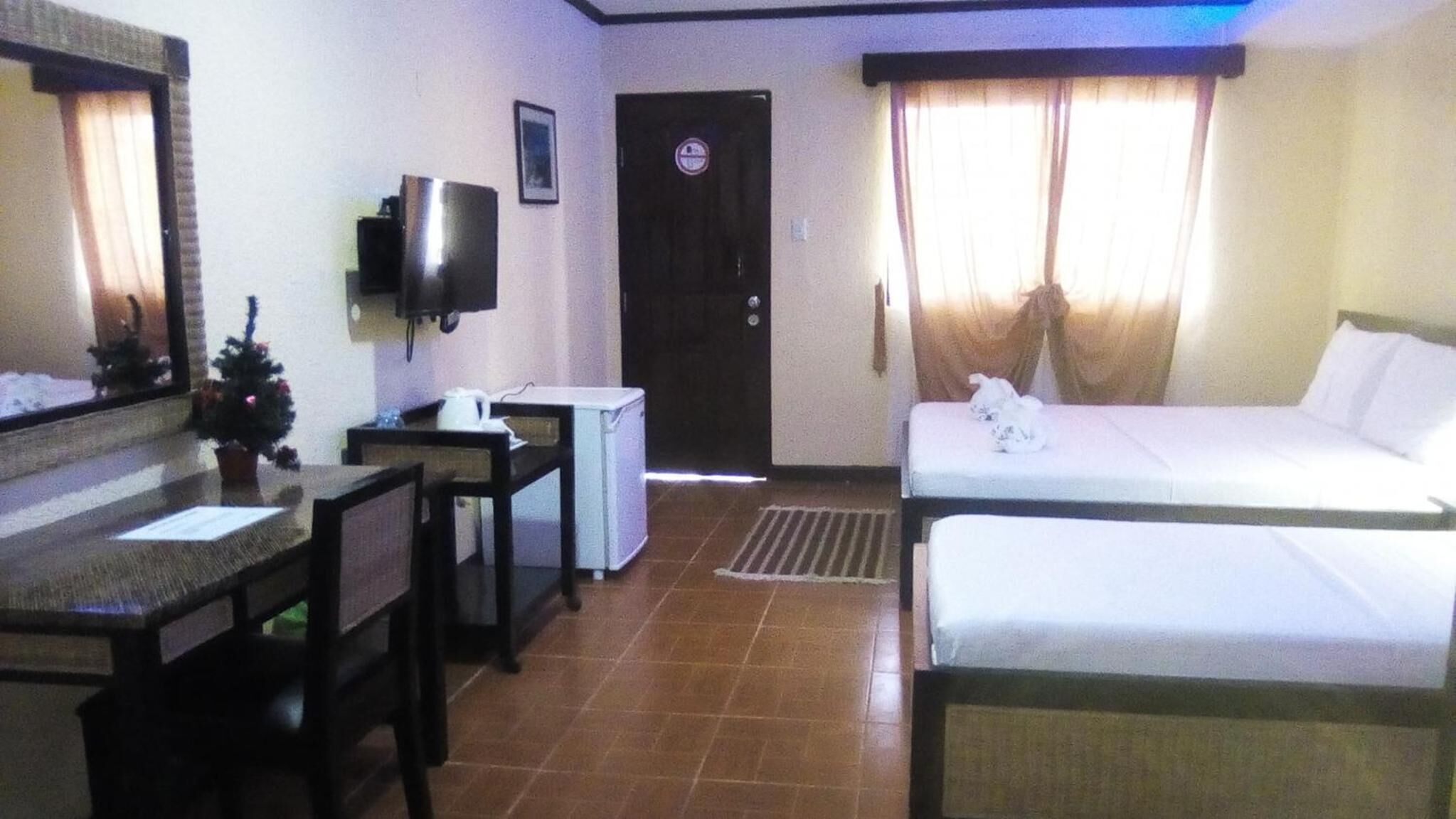 Ein Hotelzimmer mit Bett, Schreibtisch mit Stuhl, Fernseher, kleinem Kühlschrank, einer Tür mit 'No Entry'-Schild und einem Vorhangfenster.