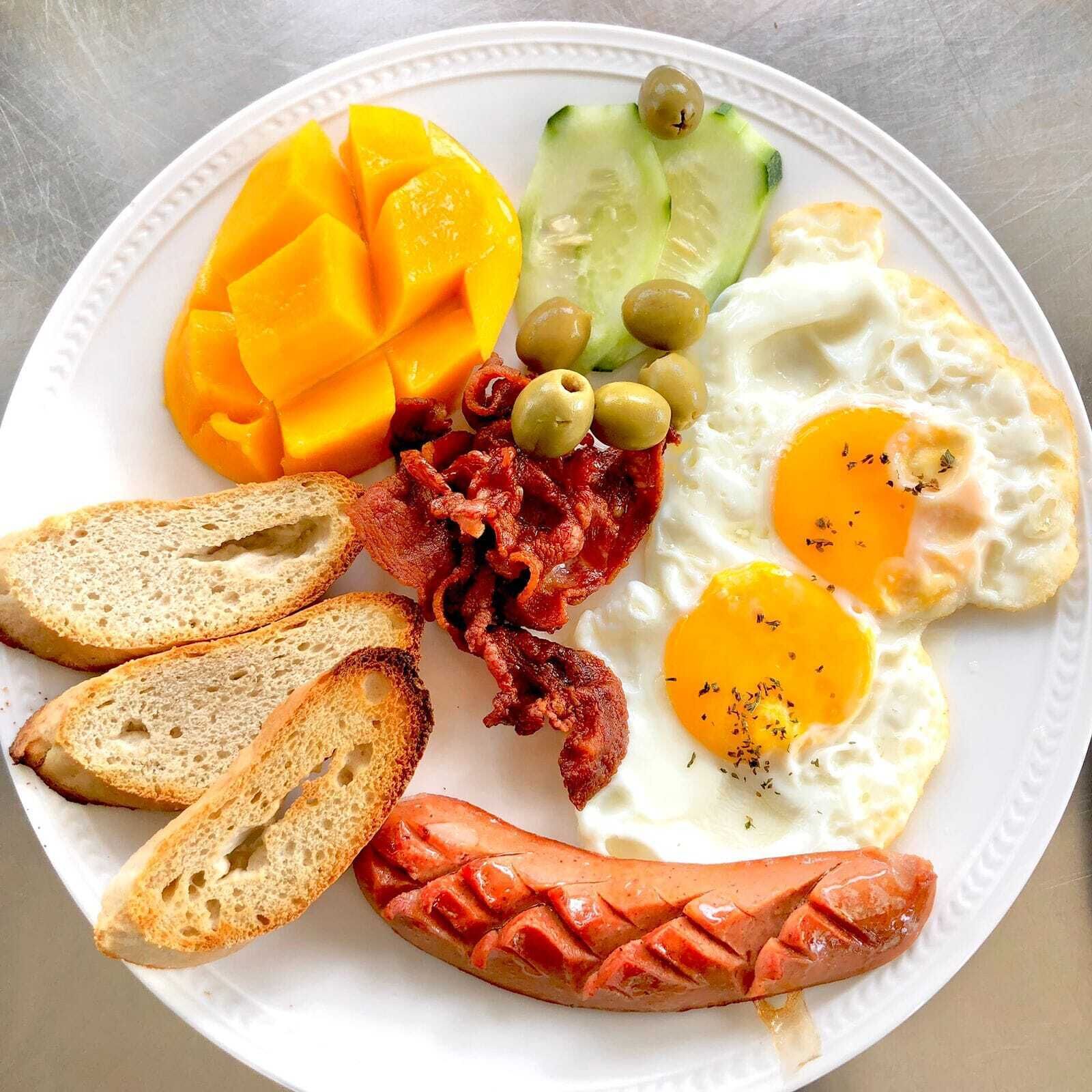 A plate with eggs, sausage, toast, mango, cucumber, and olives.