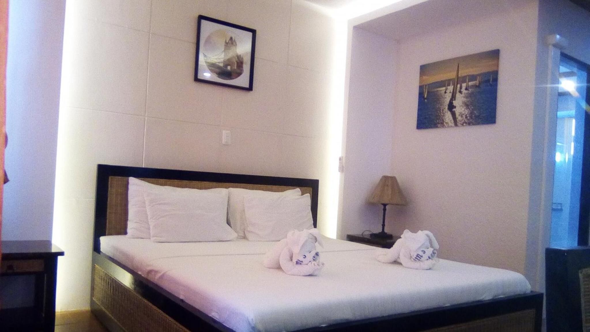 A hotel room with a bed, two pillows, a nightstand with a lamp, and two towels on the bed.