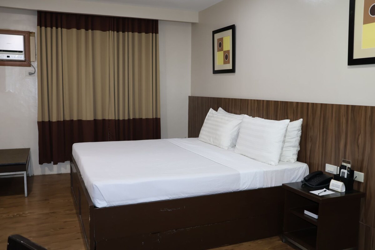 Ein Hotelzimmer mit einem großen Bett, einem Nachttisch mit Telefon und Lampe, einem Fenster mit Vorhängen und einem gerahmten Bild an der Wand.