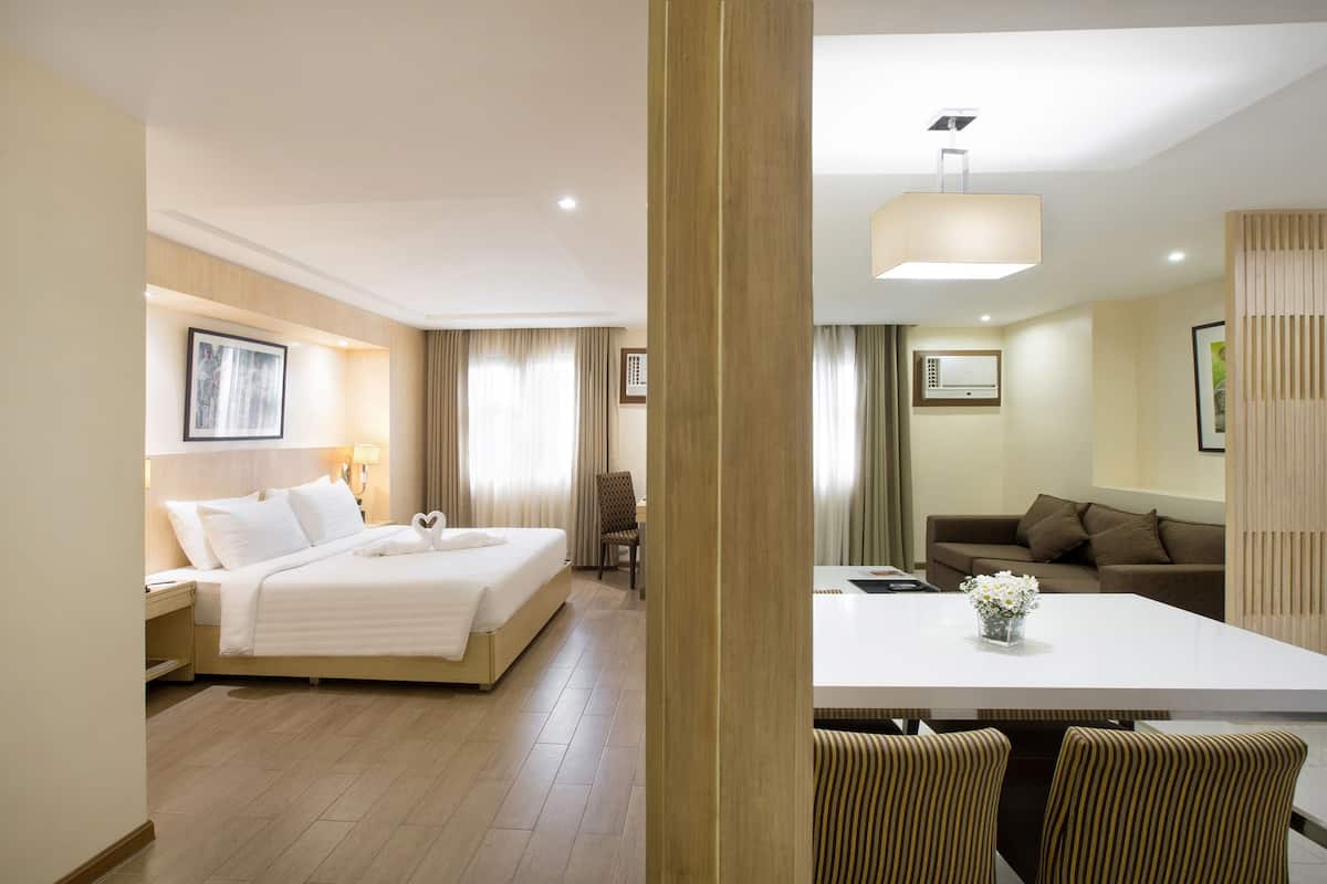 Habitación de hotel con cama, mesita de noche, lámpara, cortinas, una mesa de comedor con sillas y un sofá.