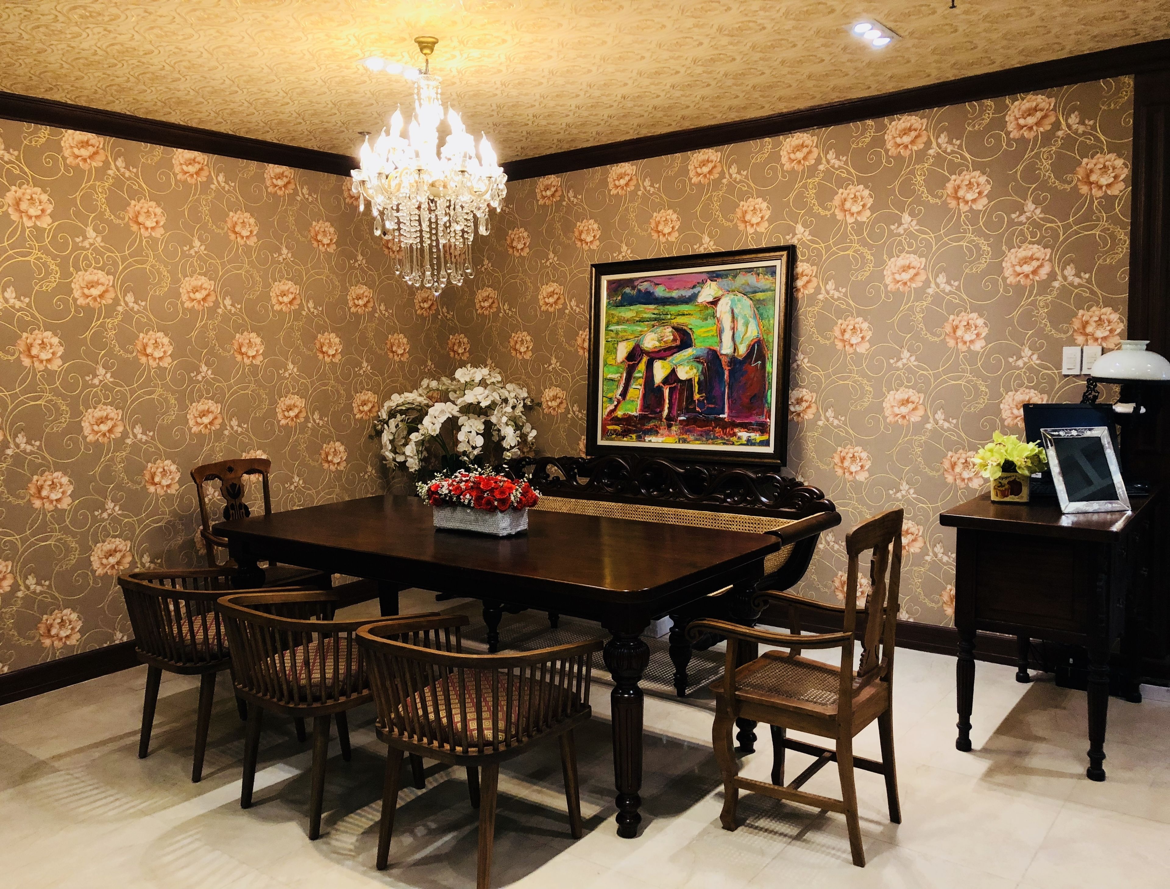 Un comedor con una mesa de madera oscura, seis sillas, una araña, papel tapiz floral y un cuadro enmarcado.