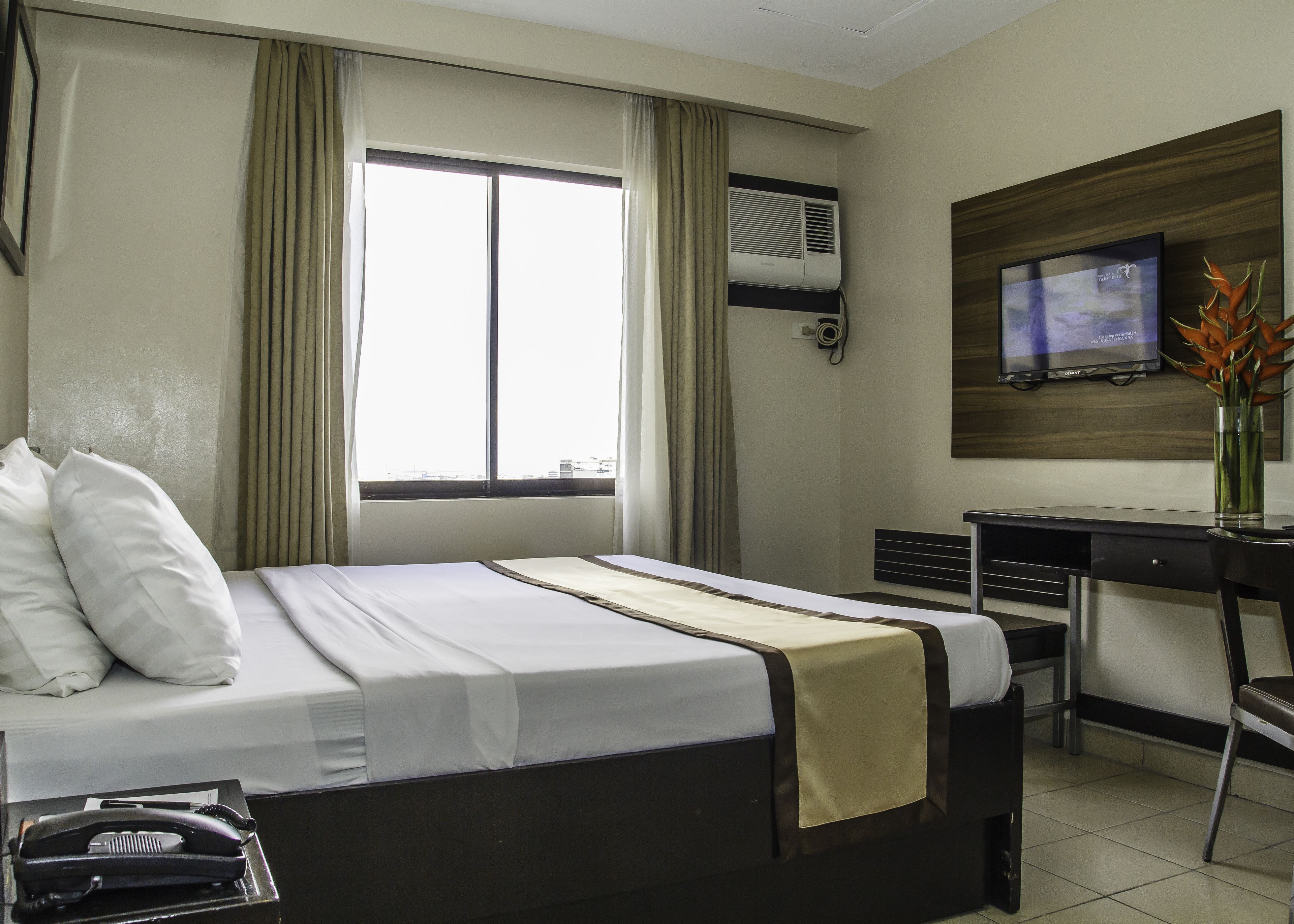 A hotel room with a bed, a desk, a chair, an air conditioning unit, a television, and a vase with flowers.