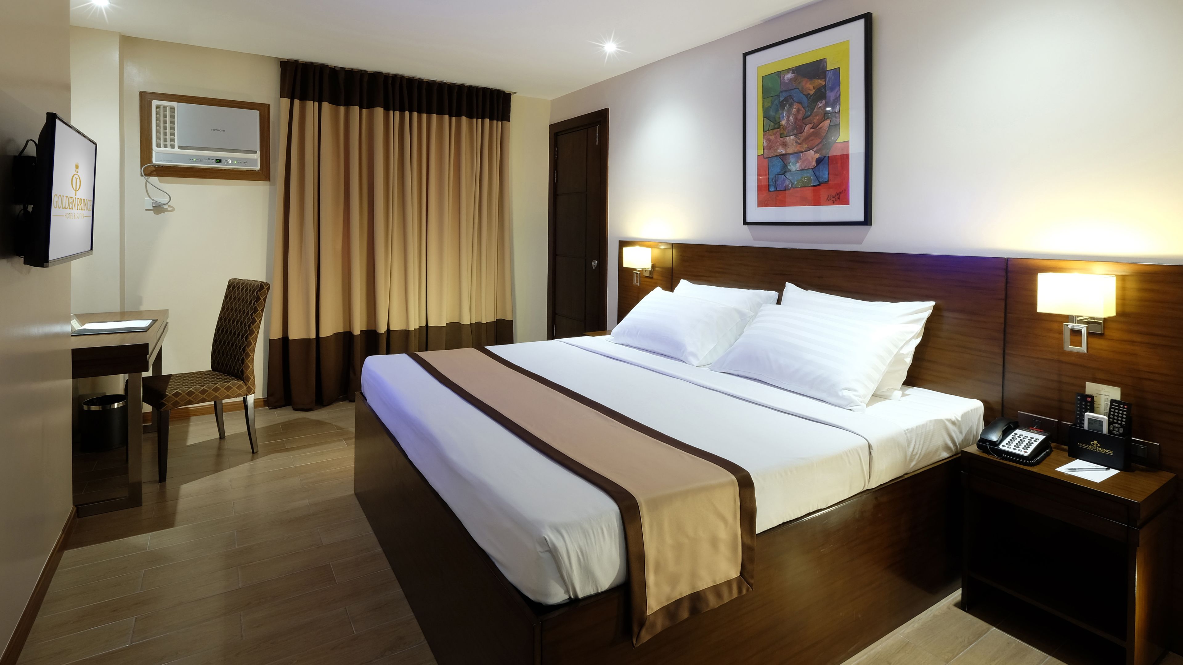 A hotel room with a bed, bedside lamps, a desk, a chair, a TV, and a painting on the wall.